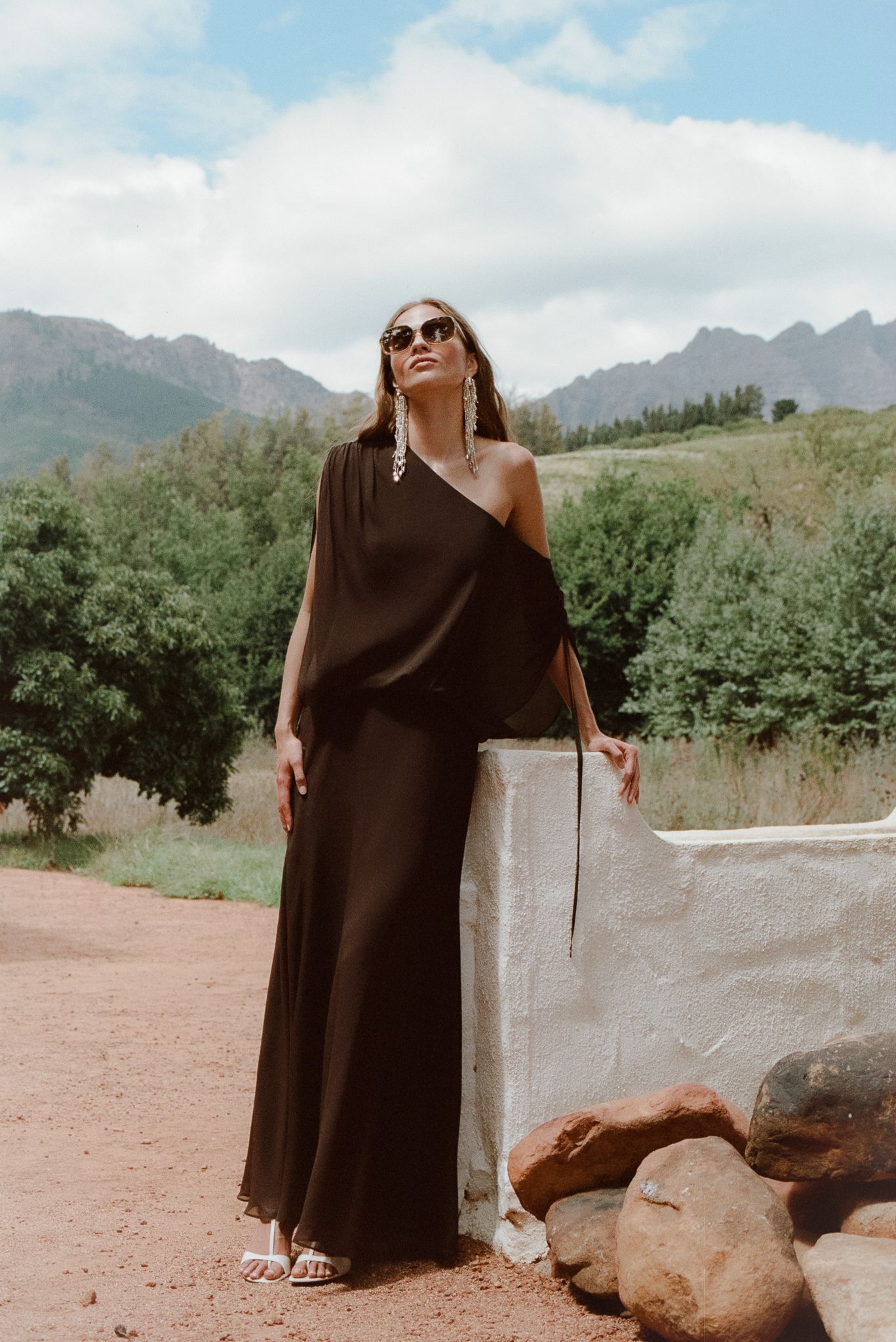Woman in black one-shoulder maxi dress and sunglasses poses outdoors with mountain backdrop.