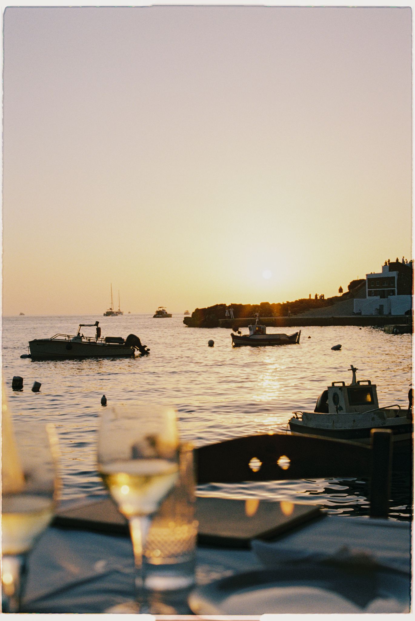 Wine glasses silhouetted against a golden sunset over a harbor with boats anchored in the tranquil waters.