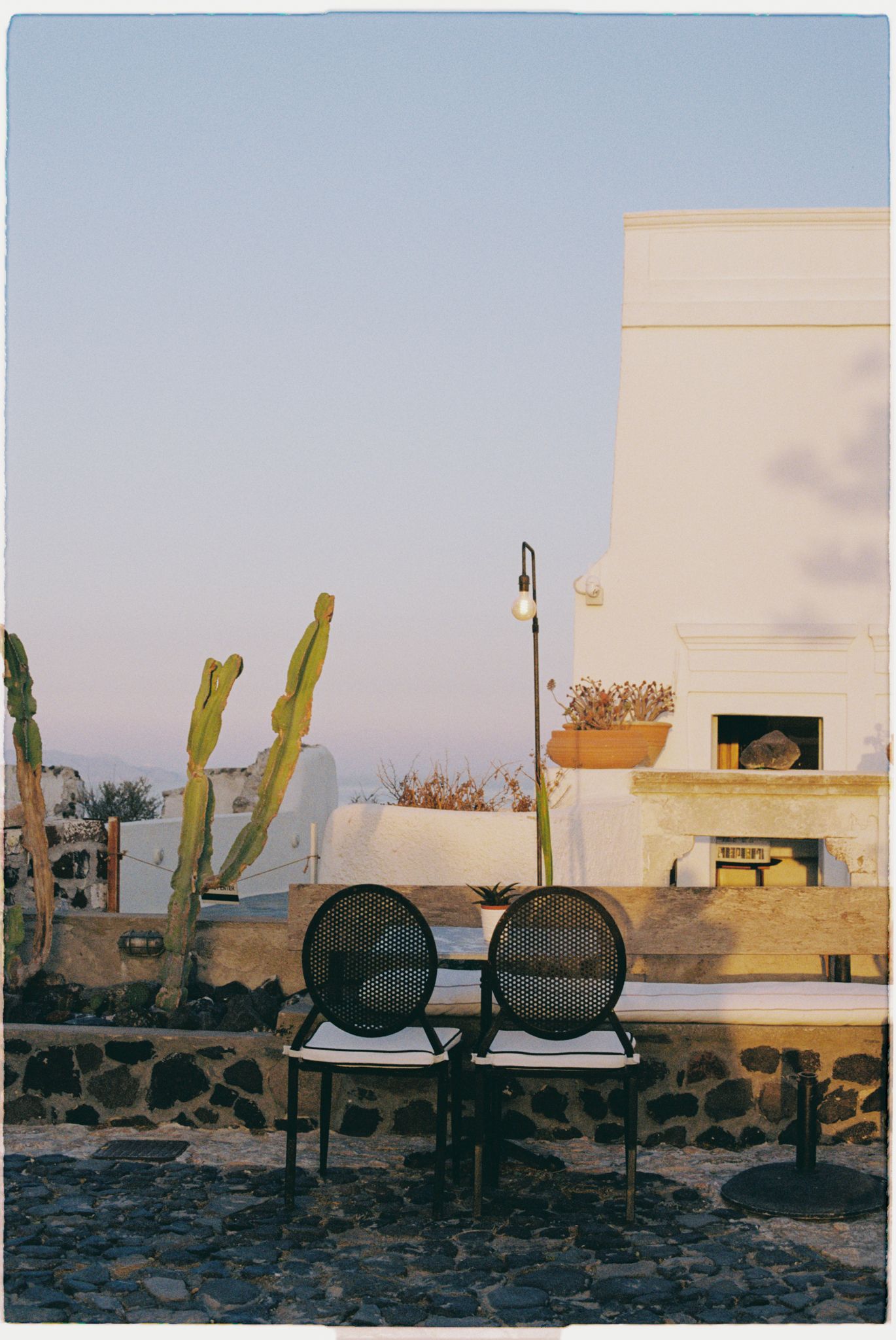 Two black round industrial drums sit on chairs against a Mediterranean-style white building with cacti at sunset.