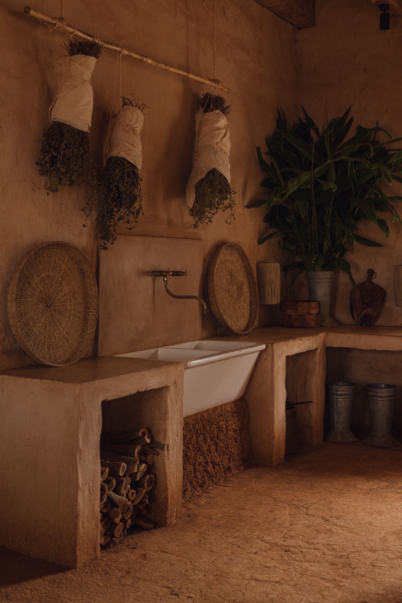 Rustic adobe kitchen with white farmhouse sink, dried herb bundles, woven baskets, and lush green plant in warm earthy tones.