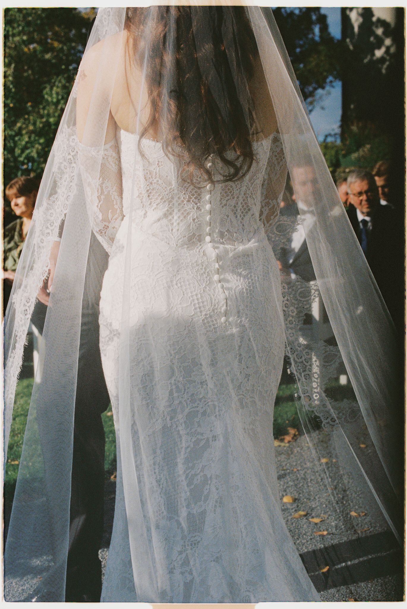 A bride in a flowing lace wedding dress and veil stands in an outdoor garden setting during a wedding ceremony.
