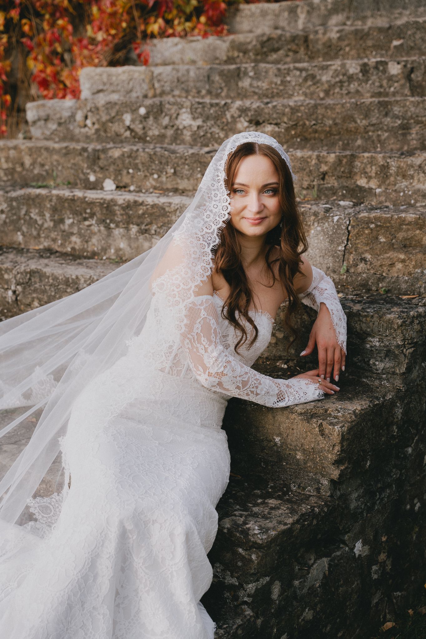A bride in a delicate lace veil poses gracefully against a weathered stone wall with autumn foliage in the background.