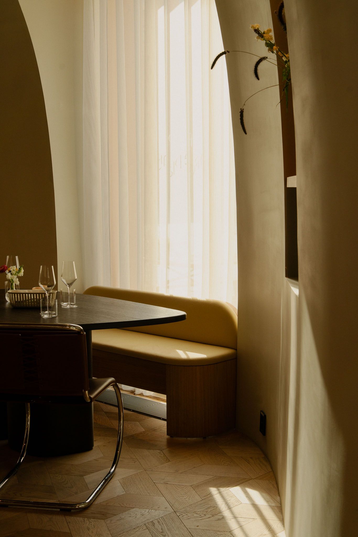 Minimalist dining nook with curved bench seating, wine glasses, and natural light streaming through vertical blinds.