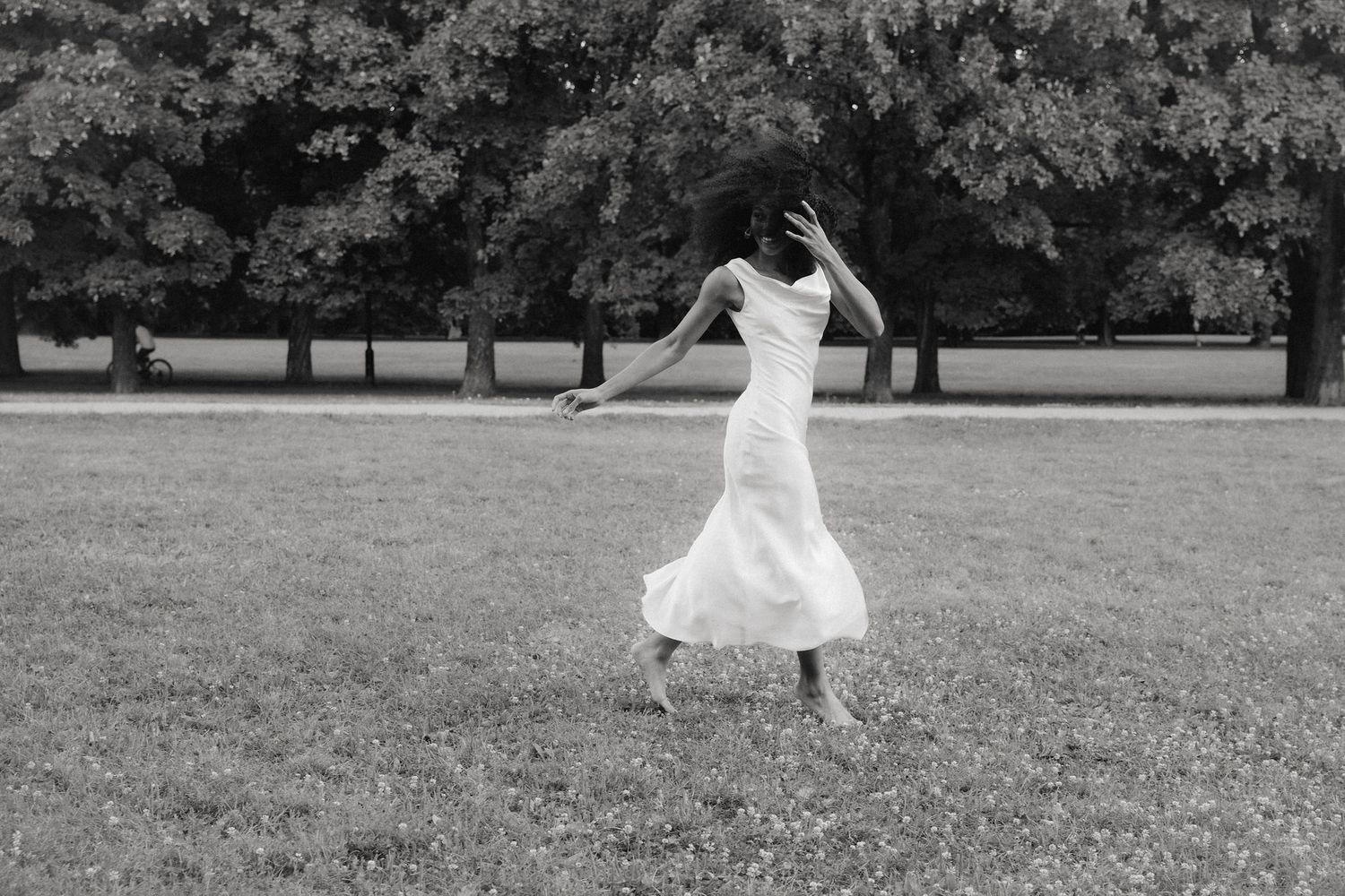 Black and white photo of a woman in a white dress dancing freely in a lush green park.
