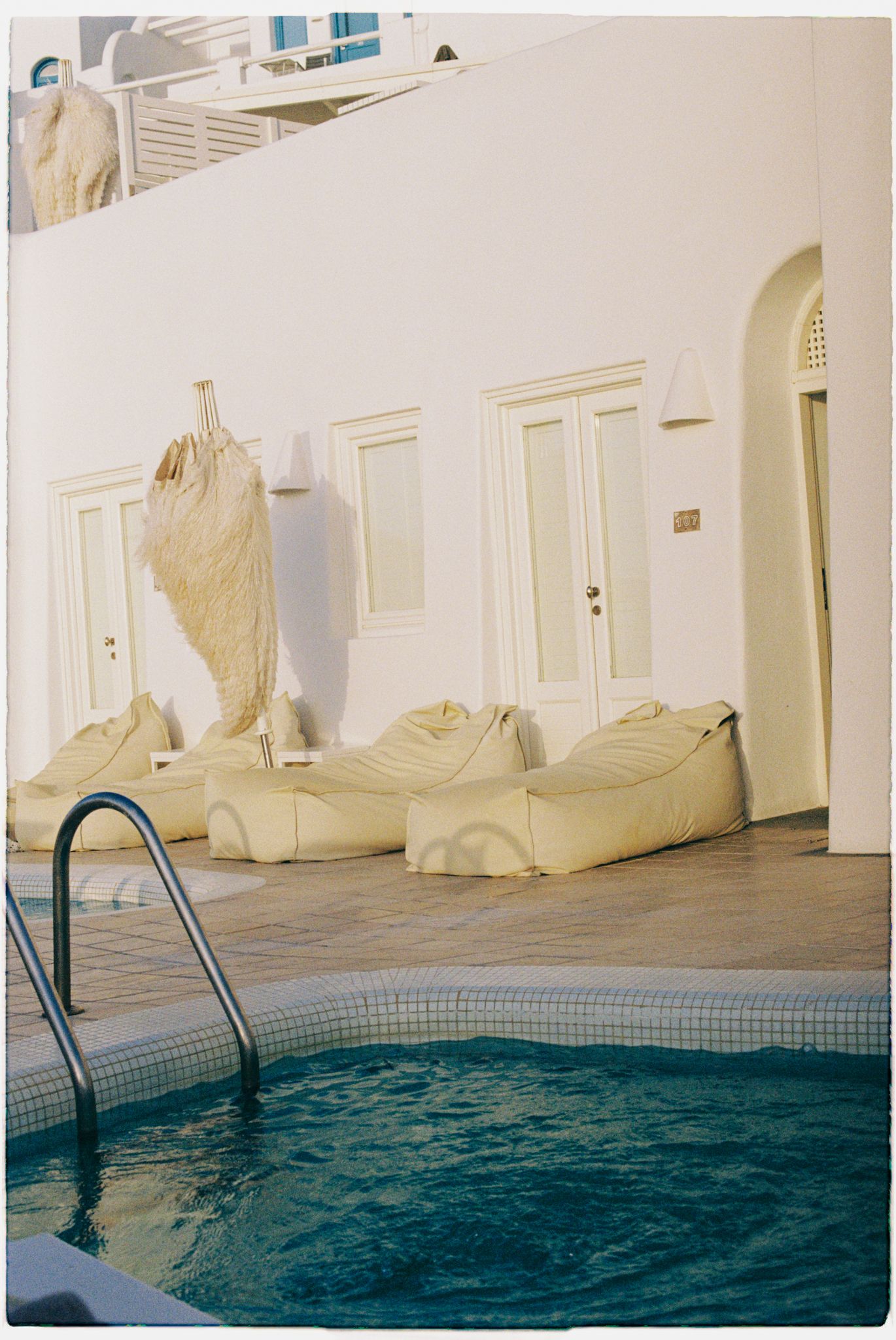Outdoor swimming pool with blue water and white walls featuring pool ladders and covered furniture during sunset.
