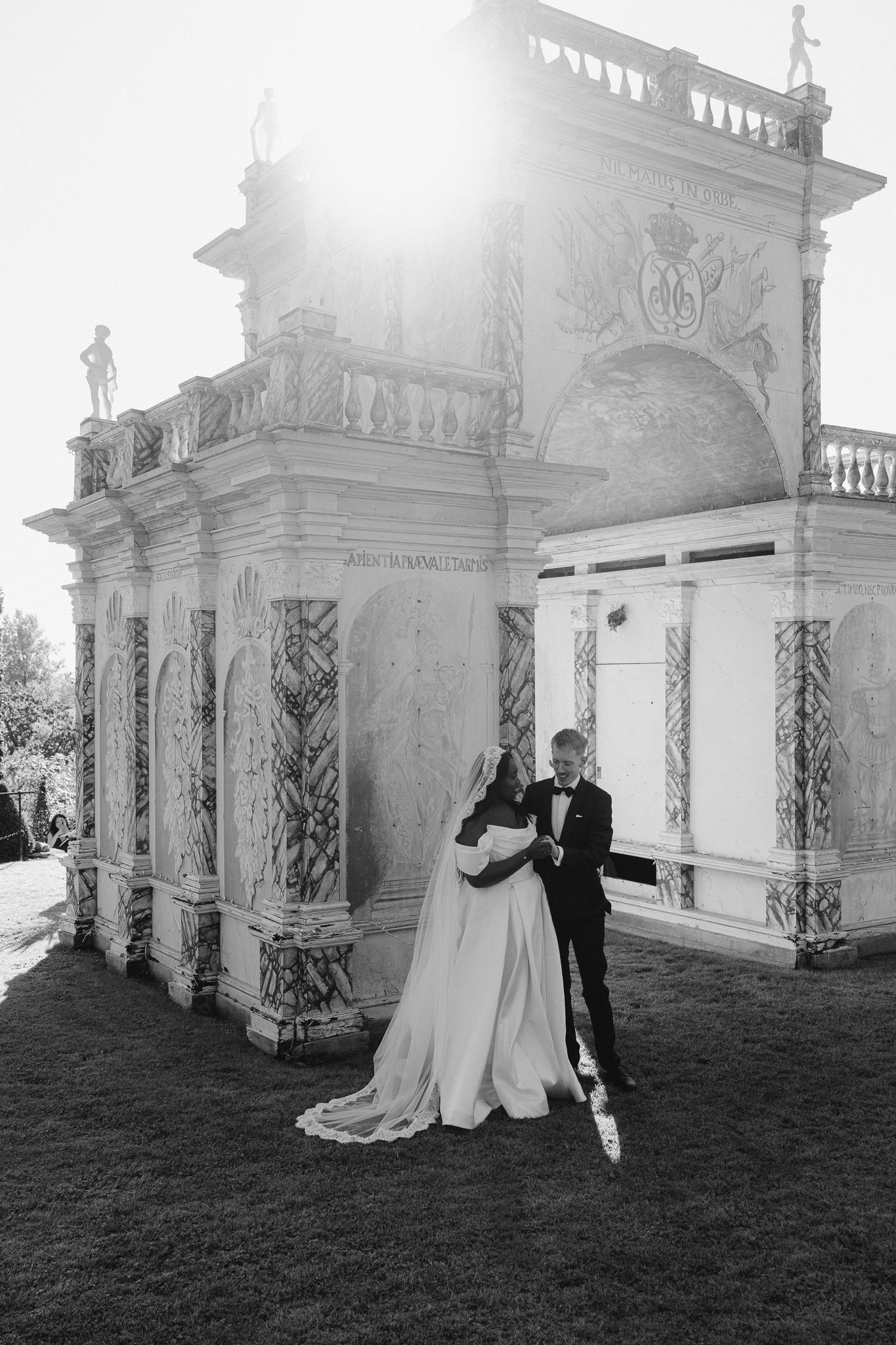 A wedding couple poses in front of an elegant white classical architectural structure in a romantic outdoor setting.