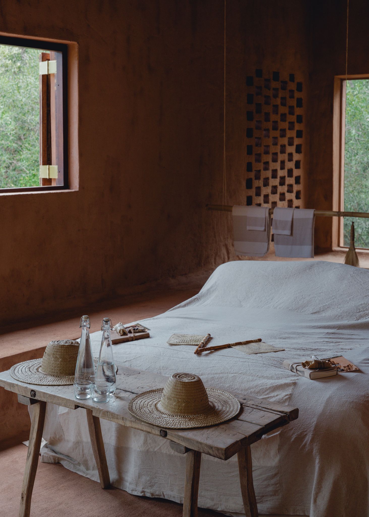 Rustic adobe bedroom with white linen bed, wooden bench, straw hats, and glass bottles near sunlit windows.