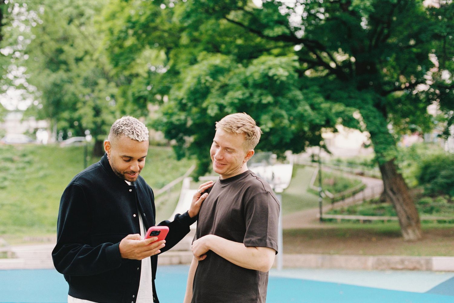 Two people share a laugh while sitting together on a park bench near a fountain on a sunny day.