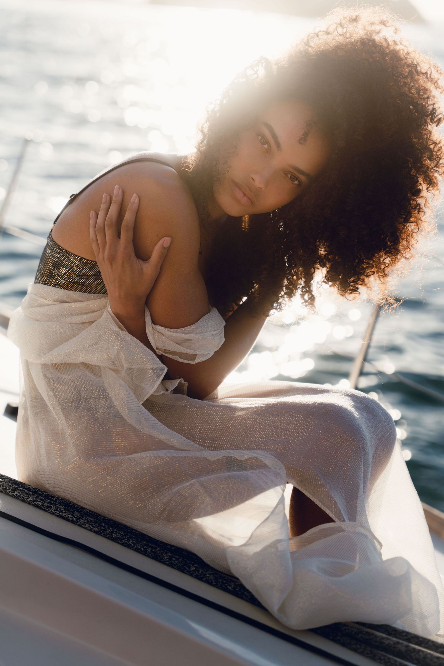 A young woman with curly hair sits gracefully on a yacht in a sheer white dress, bathed in golden sunlight over the ocean.