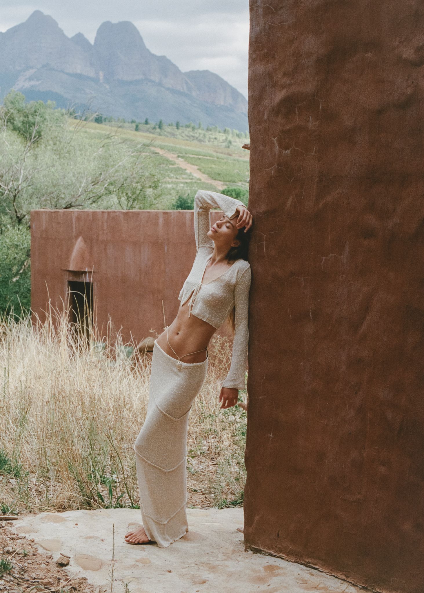 Woman in cream crop top and wide-leg pants leaning against rustic wall with mountain landscape backdrop.