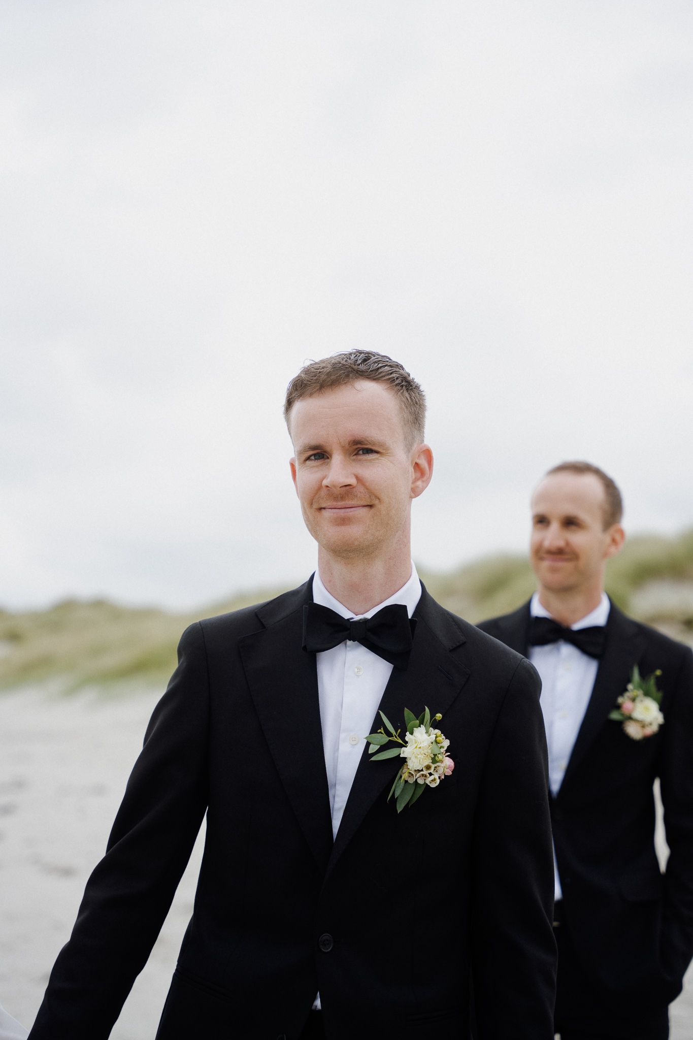 Groomsmen in black tuxedos with boutonnieres stand together on a beach during an outdoor wedding.