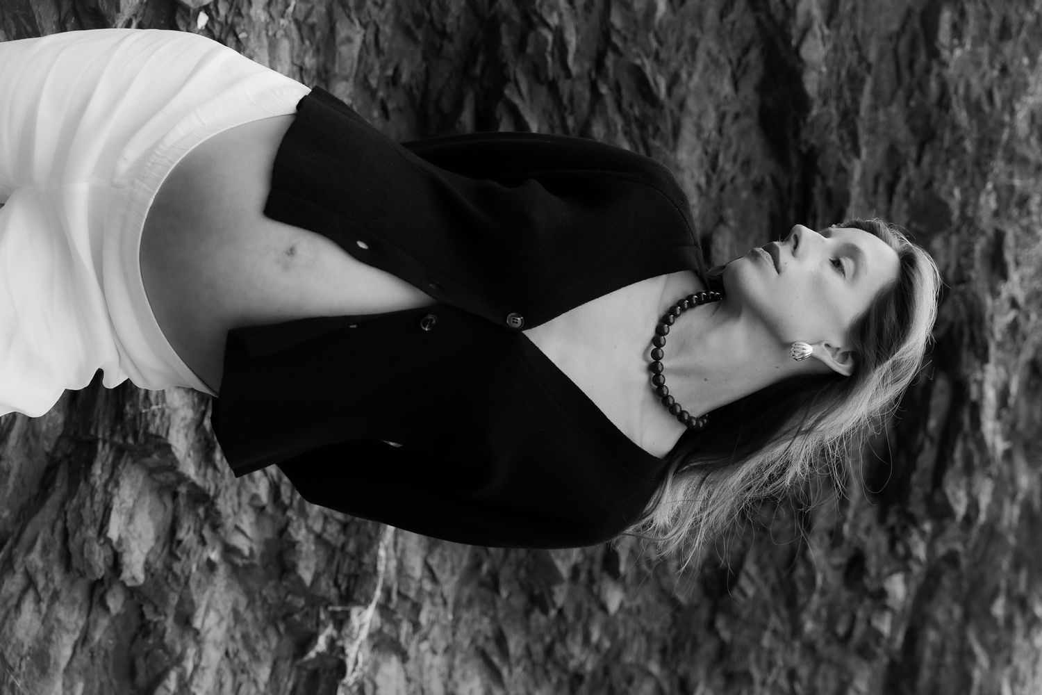 Black and white photo of a woman in a black jacket and white skirt leaning against a textured tree trunk.