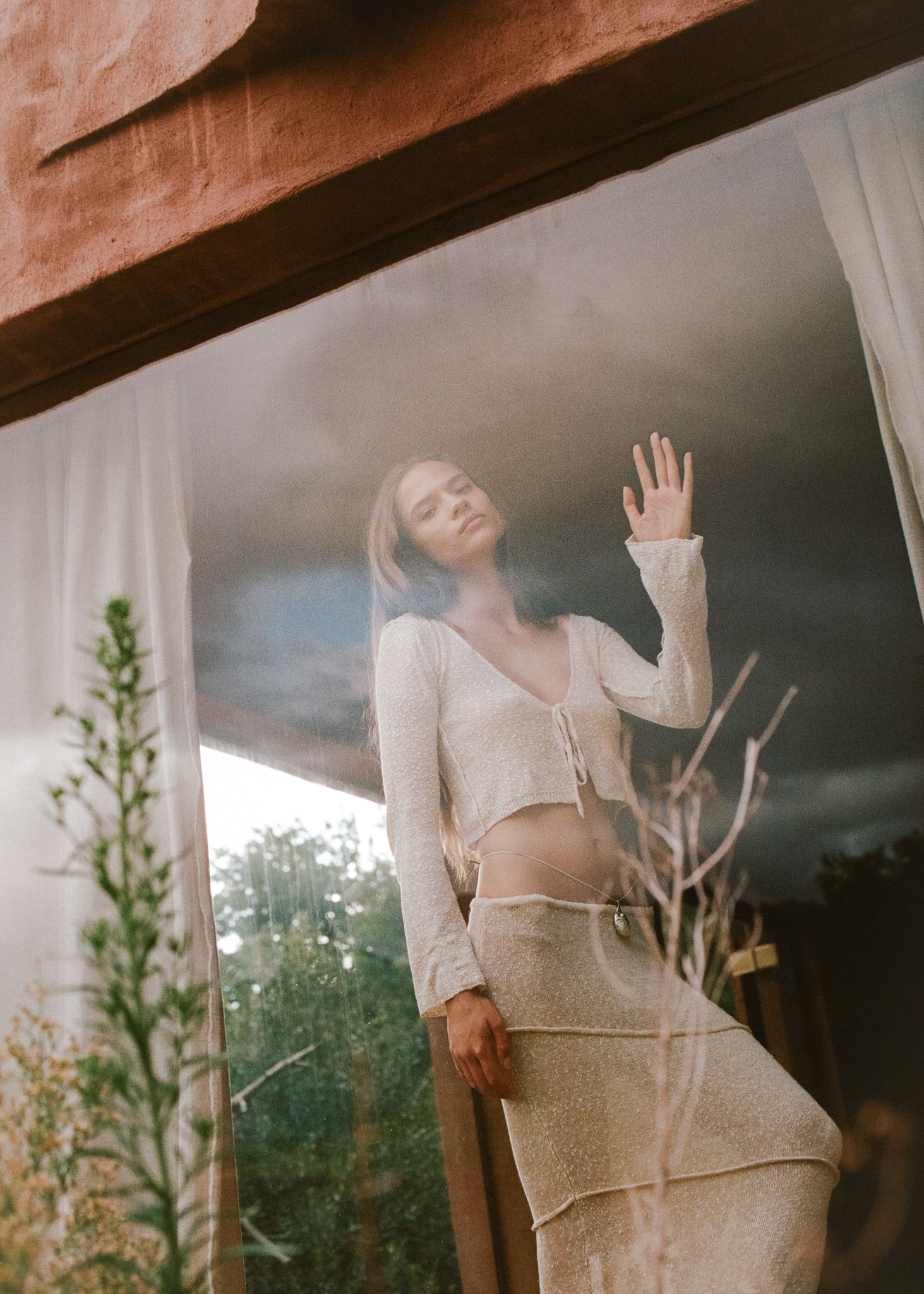 Woman in cream knit cardigan and skirt poses gracefully by a window with stormy sky backdrop.