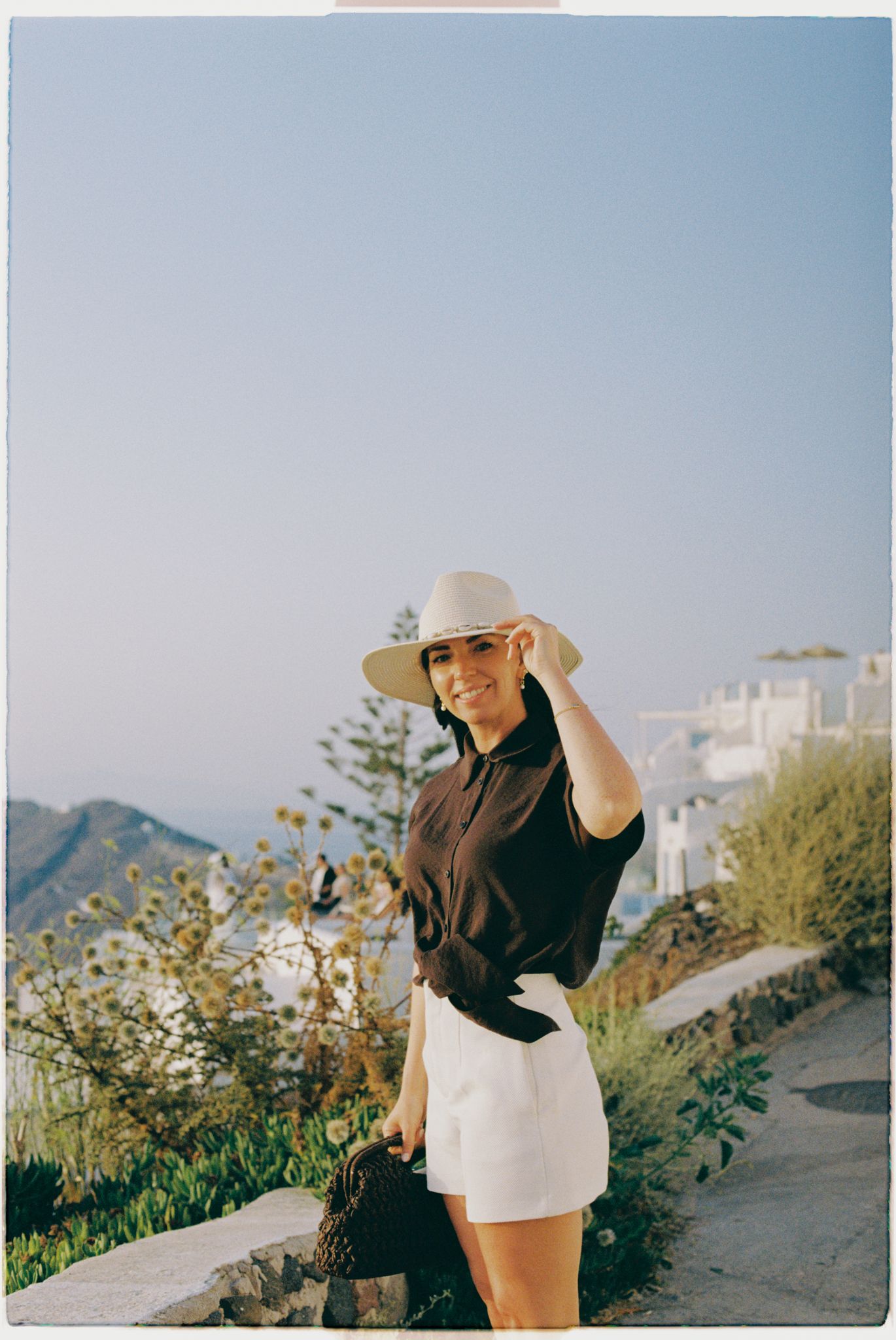 A woman n in black top and white shorts poses against a scenic hillside backdrop of Imerovigli, Santorini during golden hour.