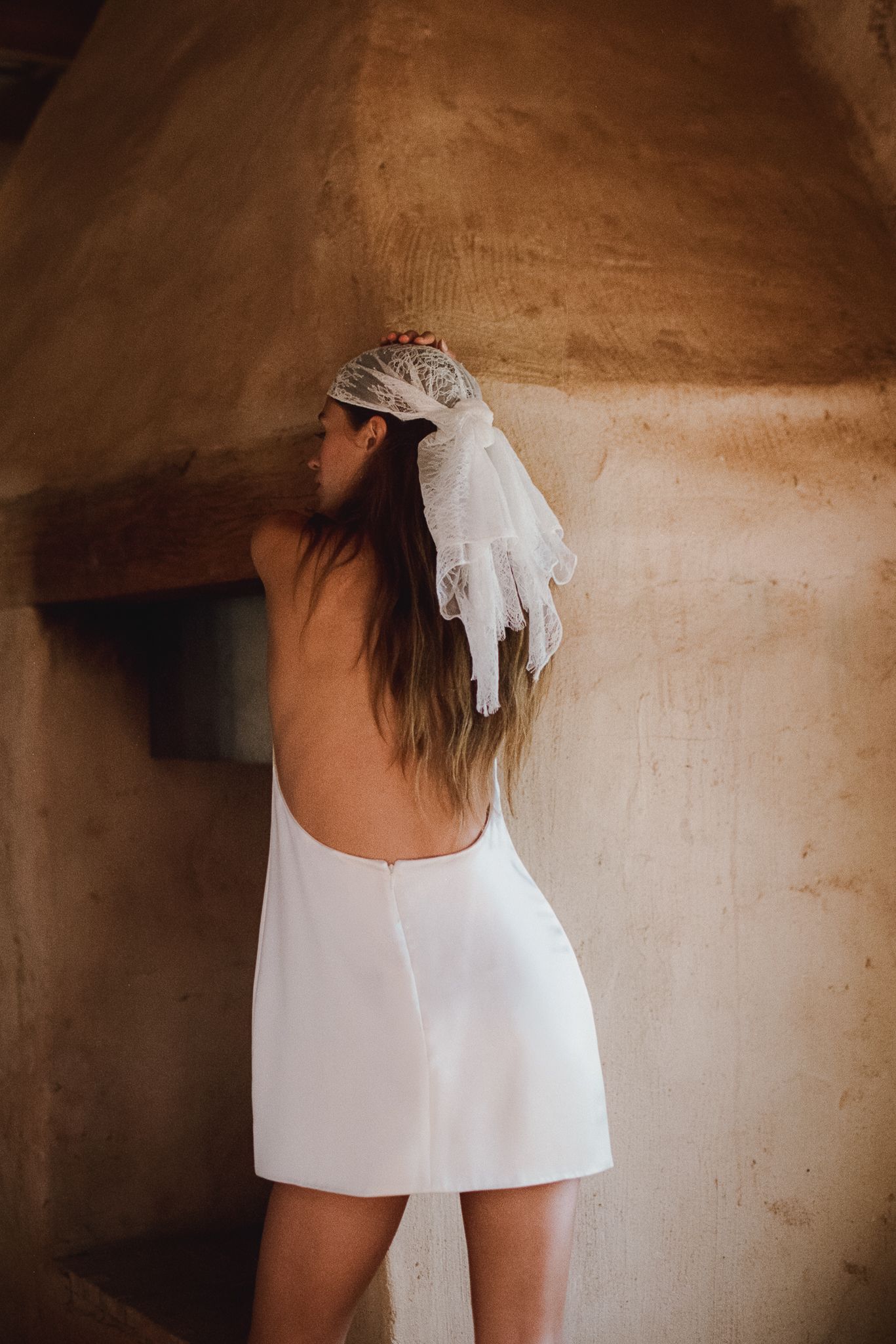 Woman in backless white mini dress with lace hair bow poses against rustic stone wall.