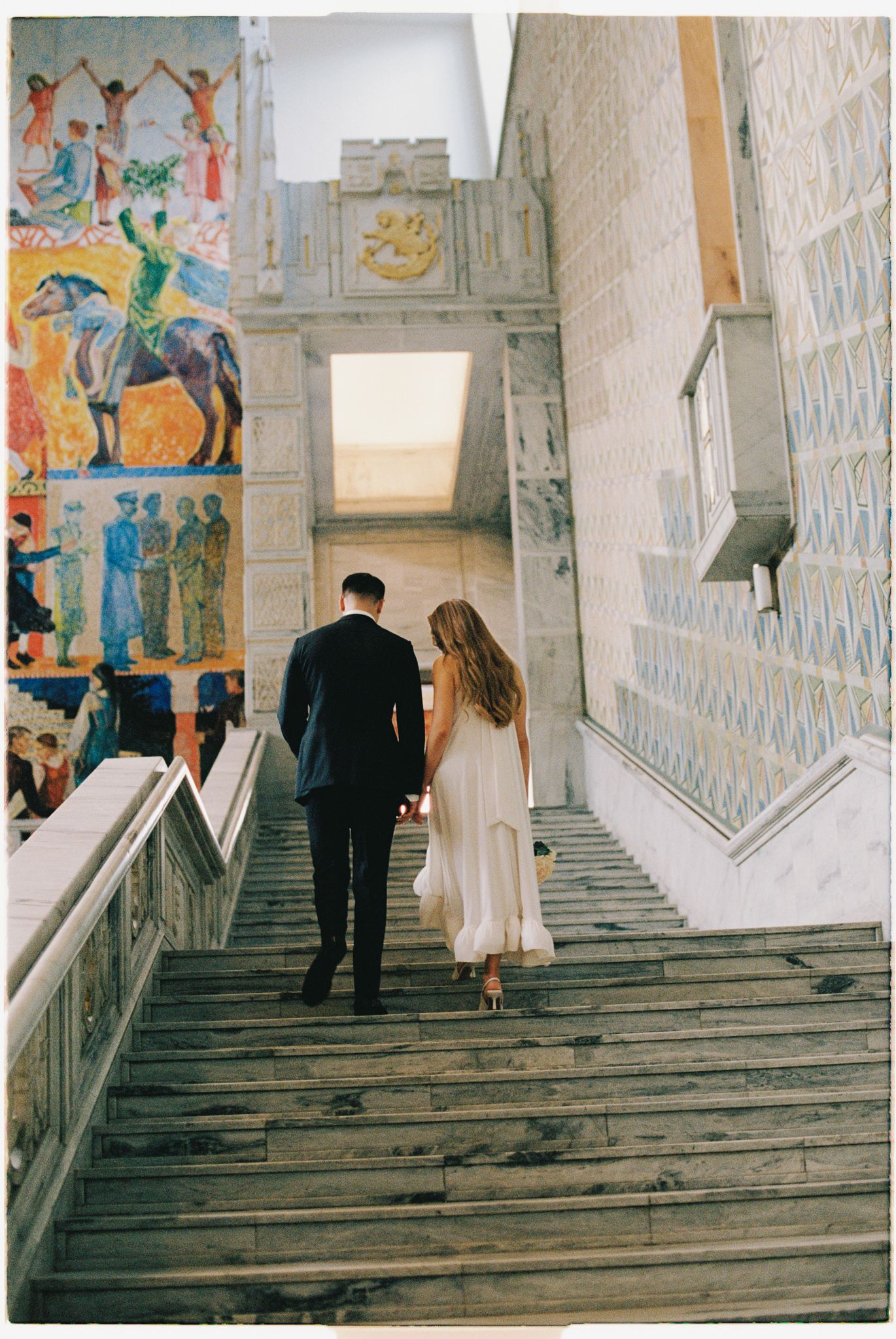 A couple ascends ornate stone stairs with decorative murals and artwork on the walls inside a historic building.