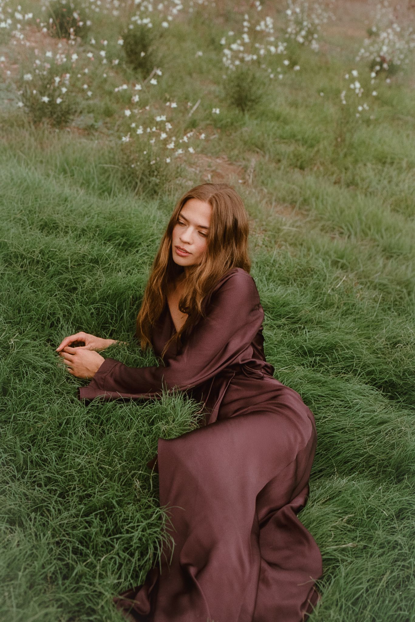Young woman in brown satin dress sitting gracefully on lush green grass field with wildflowers in background.