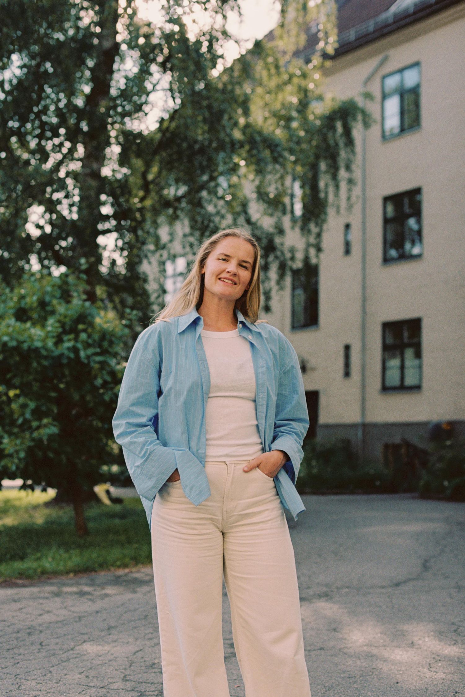 Someone wearing a denim jacket and white pants poses casually against an outdoor building backdrop at sunset.