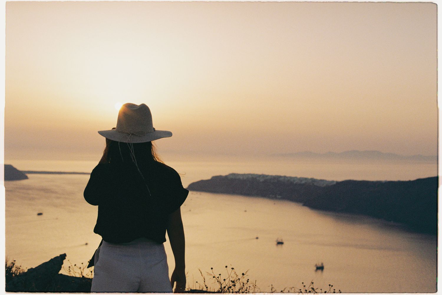A silhouetted figure admires the warm sunset over calm ocean waters and mountainous coastline views.