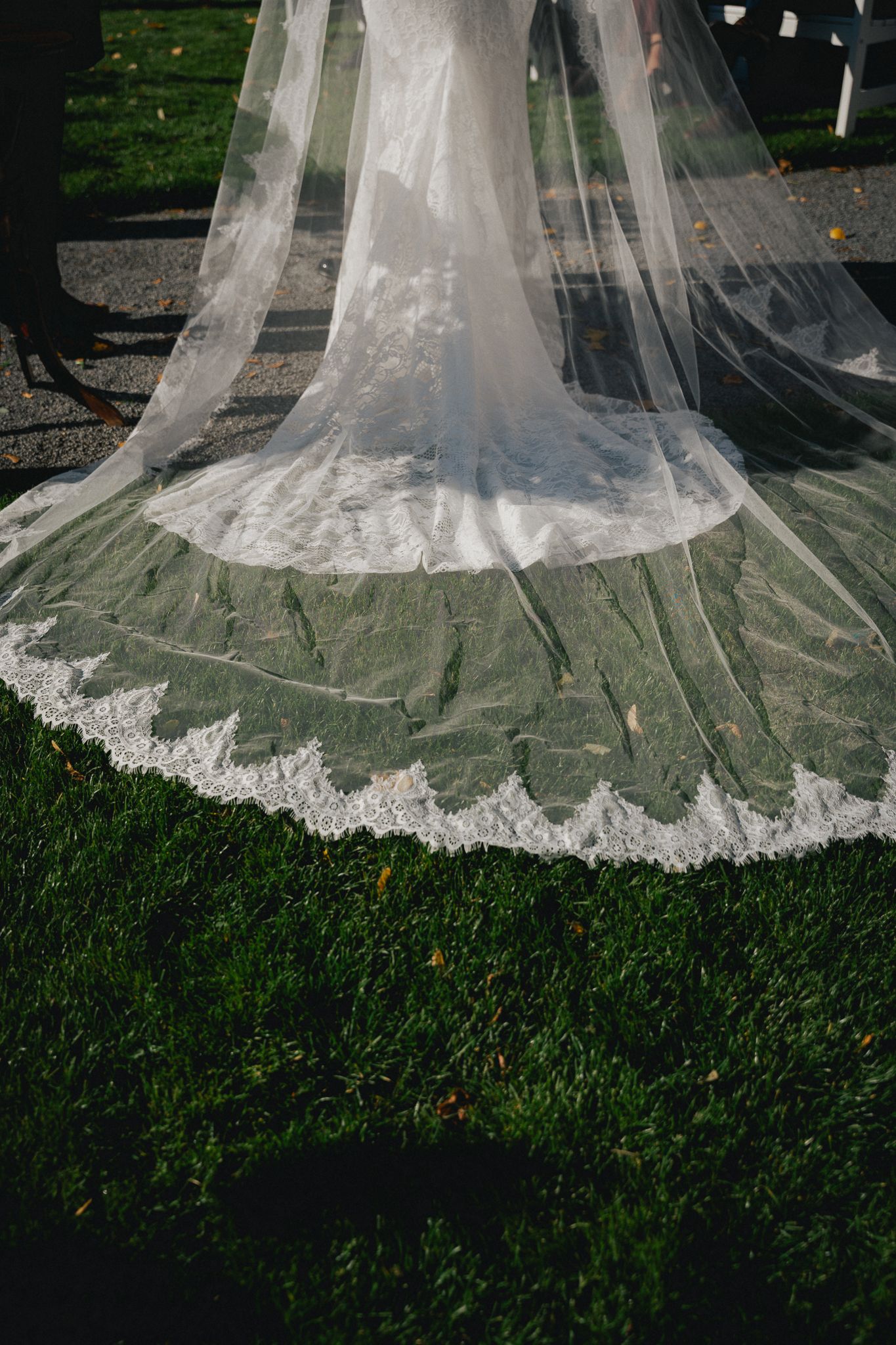 Close-up details of delicate lace trim on a white wedding dress veil trailing across green grass.