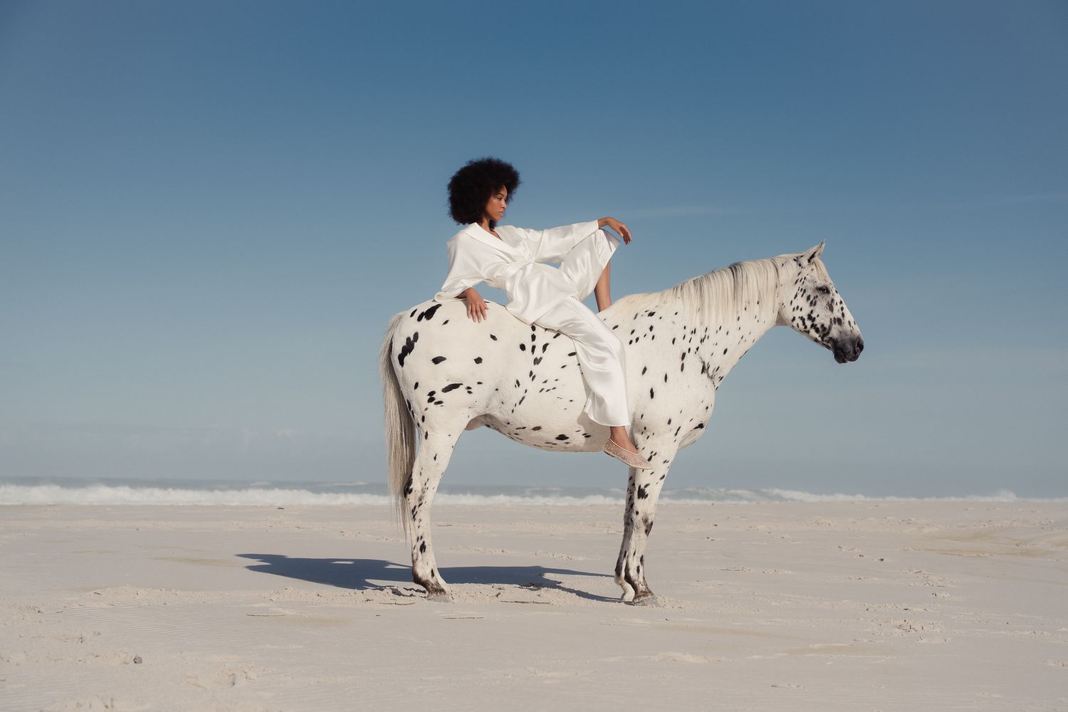 Woman in white dress riding a spotted Appaloosa horse on a sandy beach under a clear blue sky.