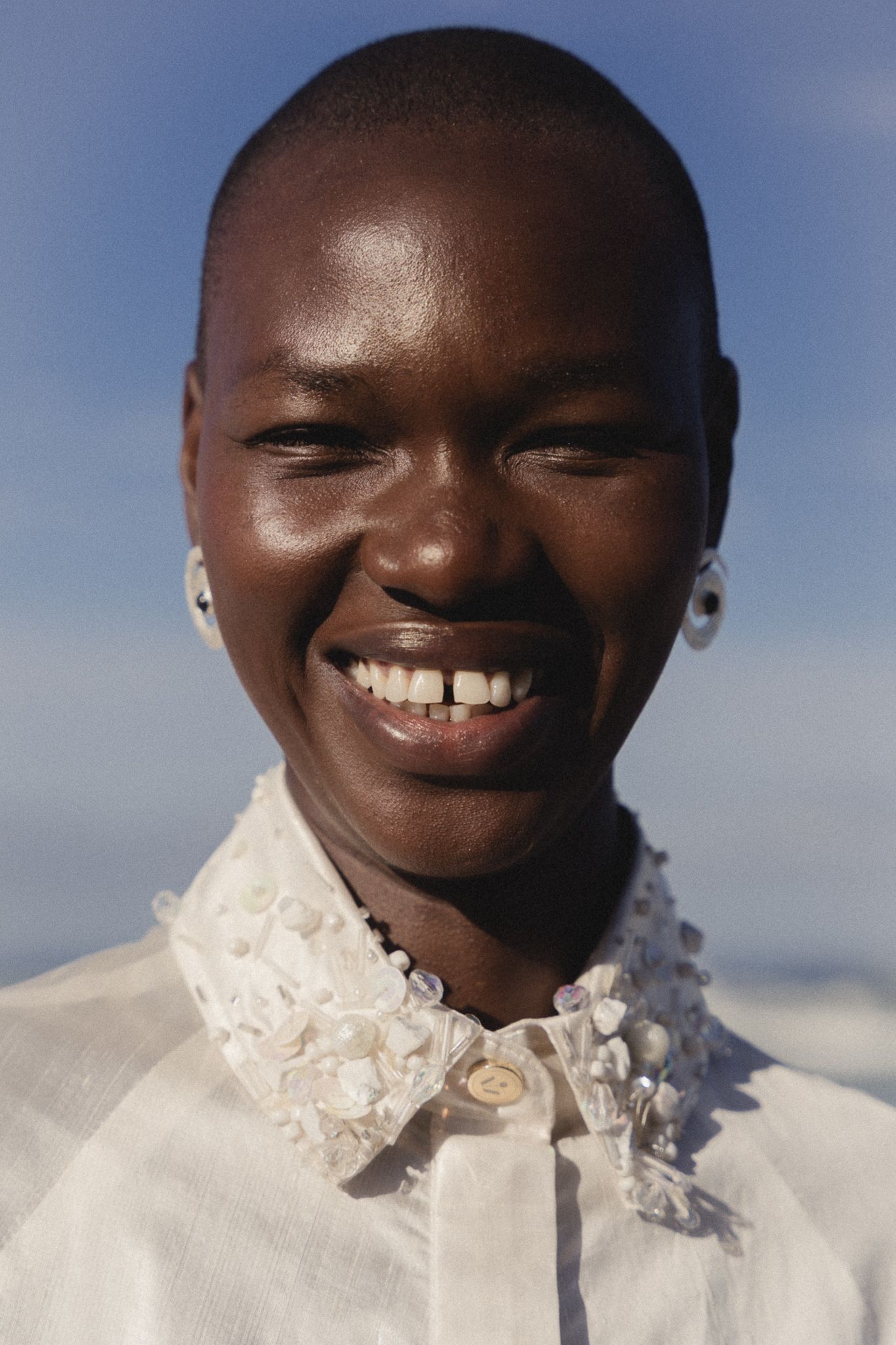 Smiling person with shaved head wearing a white embroidered collar shirt against a blue sky backdrop.