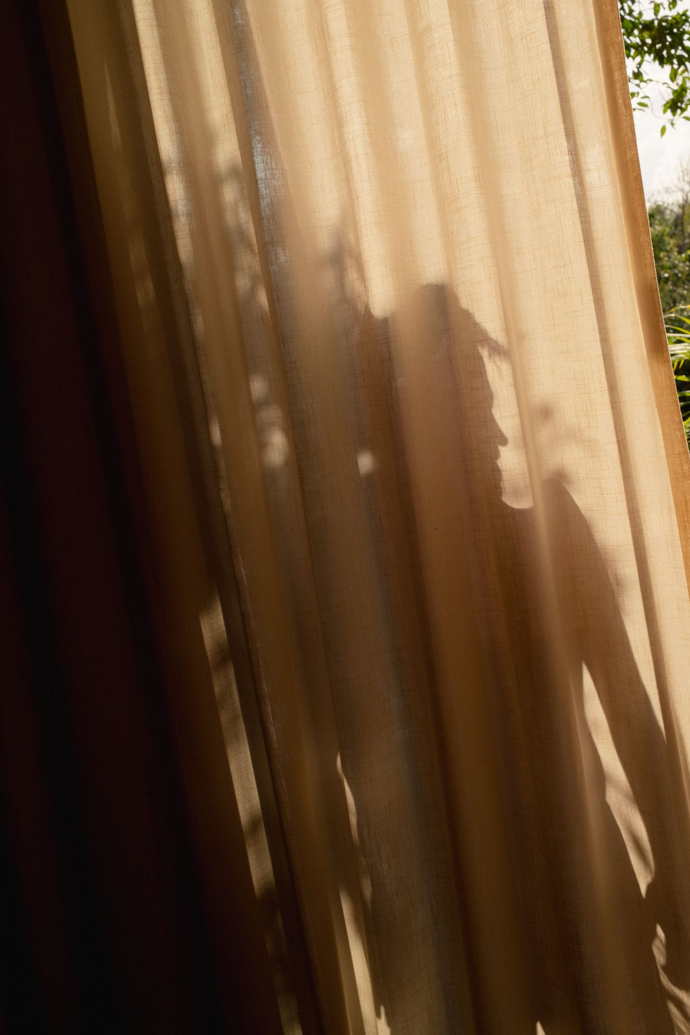 Sunlight casts dramatic shadows through sheer curtains creating an ethereal pattern on a beige wall in the apartment of Aflora, Tulum.
