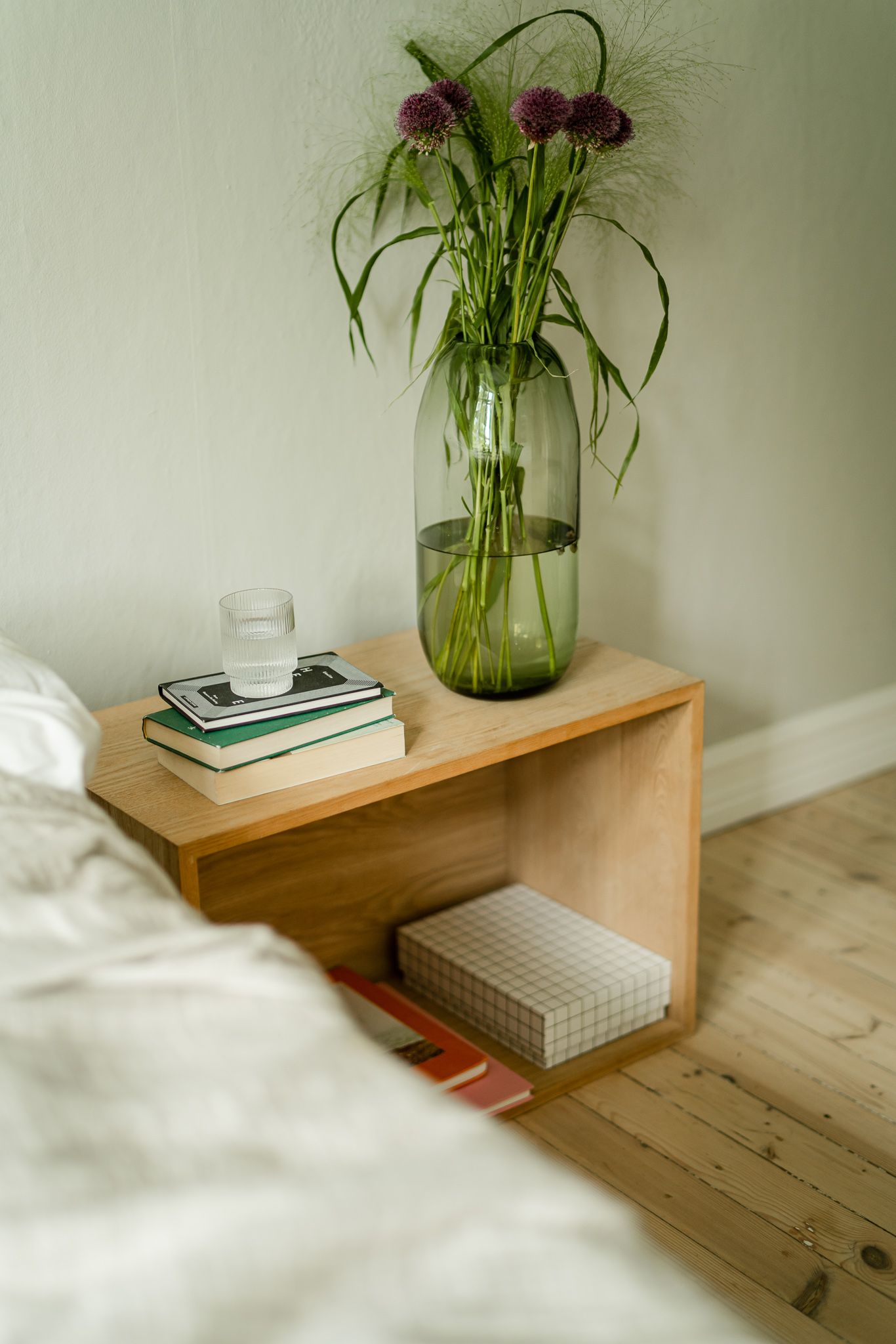 Minimalist wooden shelf with books and flowers in vase against light green wall.