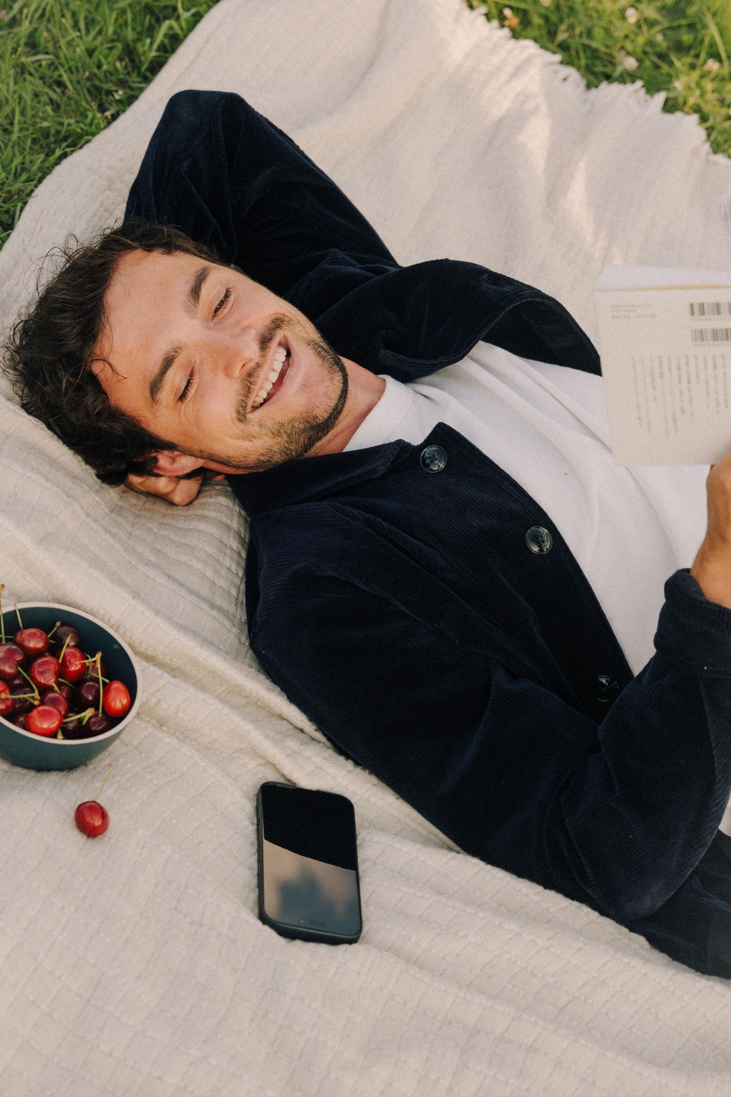 Person in black sweater relaxing on white blanket with bowl of strawberries and phone nearby.