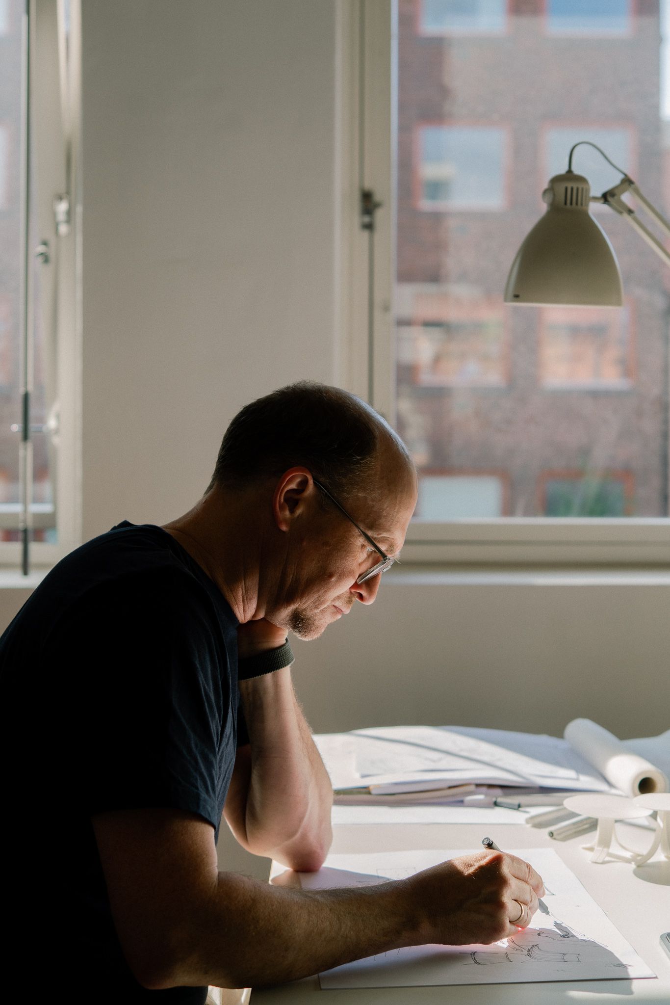 A person in a dark shirt works intently at a desk with papers and books under natural window lighting.
