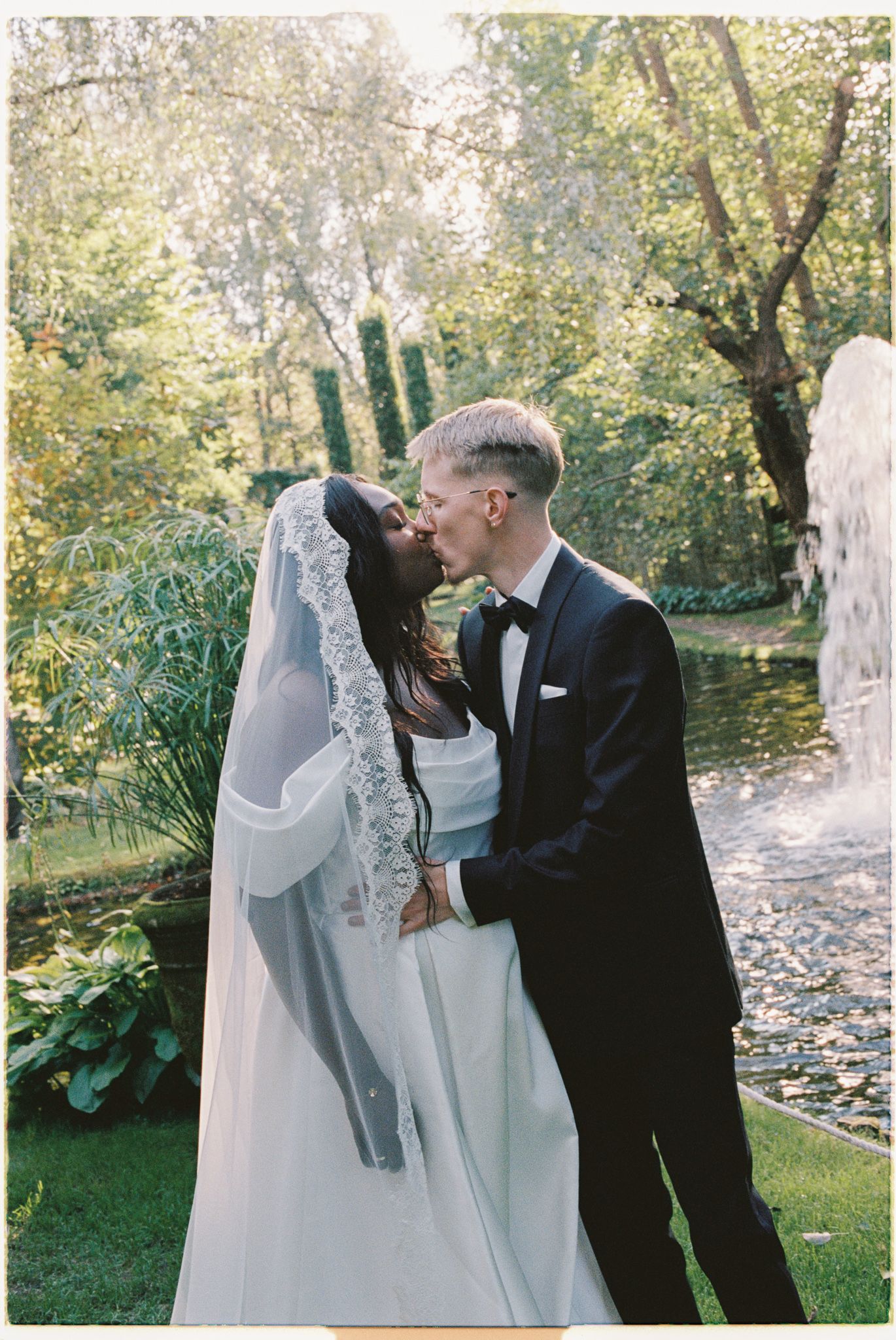 A couple shares a romantic kiss in a garden setting with autumn leaves scattered on the ground.