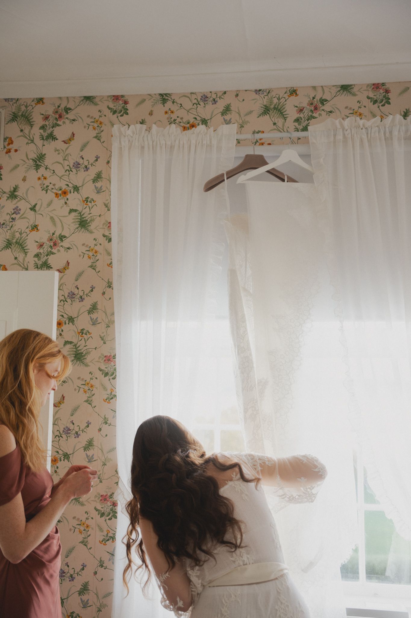 A person in a light-colored top hangs sheer white curtains on a window with floral wallpaper in the background.