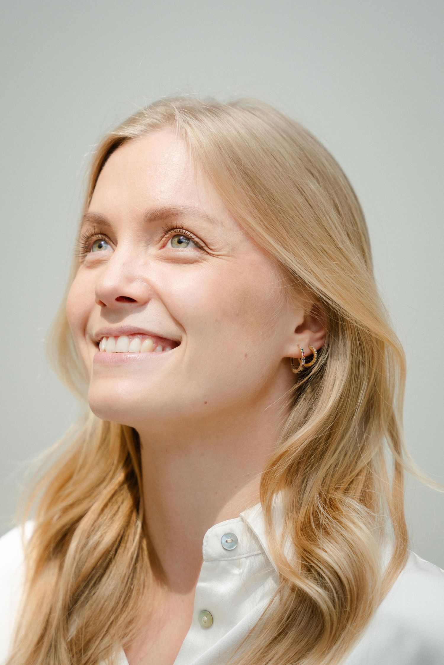 Portrait of a smiling person with long blonde wavy hair wearing a white button-up shirt against a light gray background.