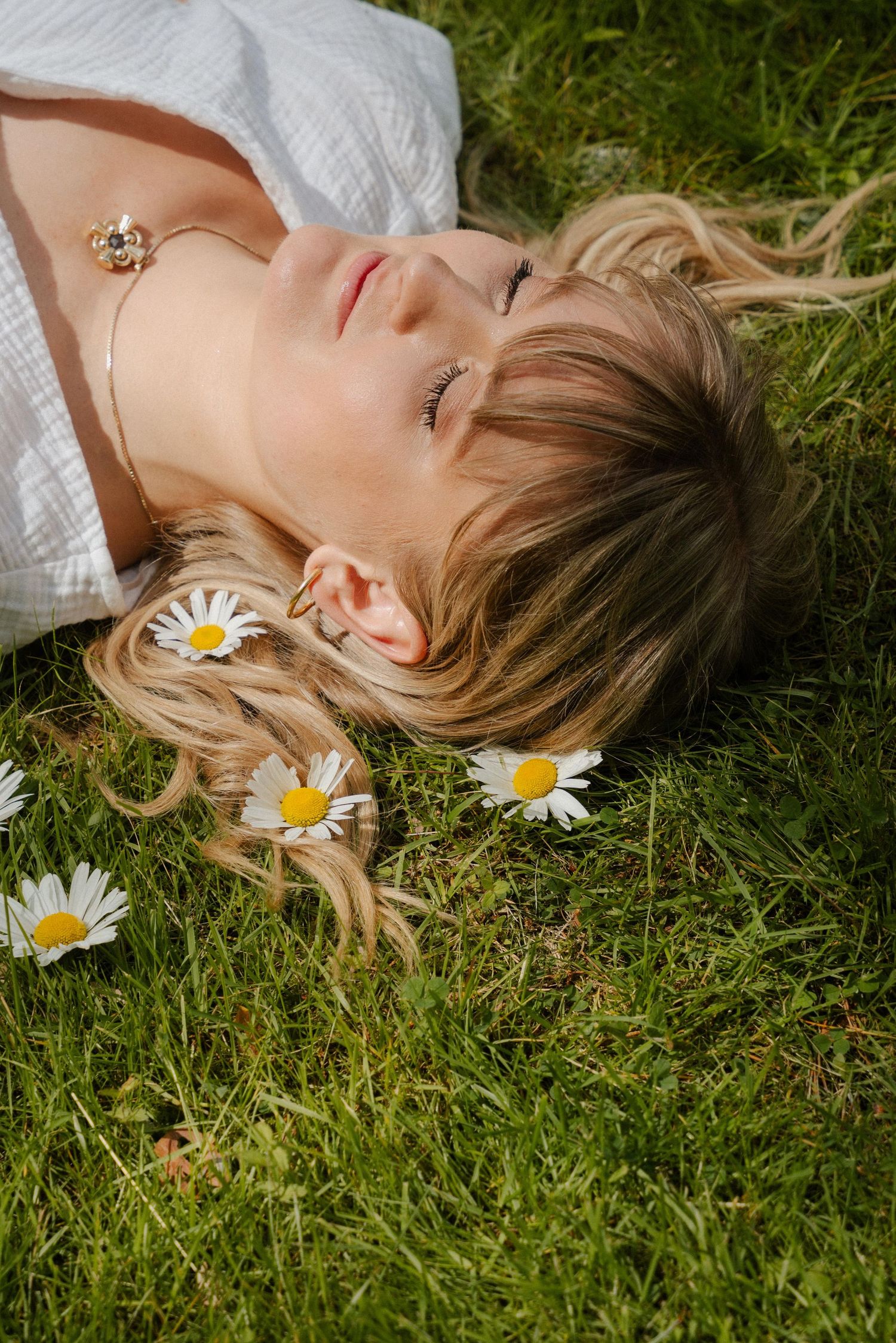 A dreamy summer portrait features someone lying in green grass with white daisies creating a whimsical natural setting.