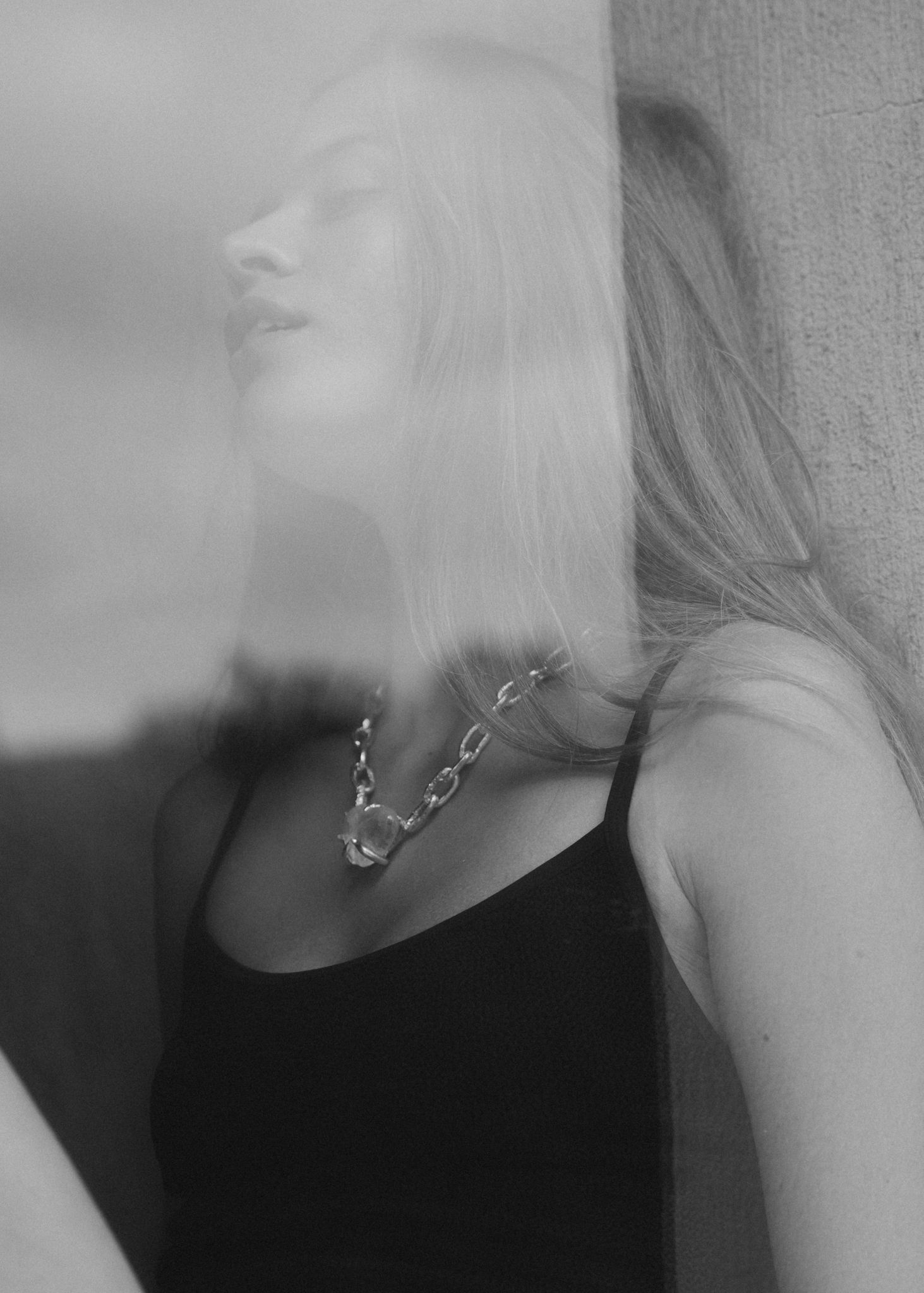 Black and white double exposure photo of blonde woman wearing chain necklace and black top leaning against wall.