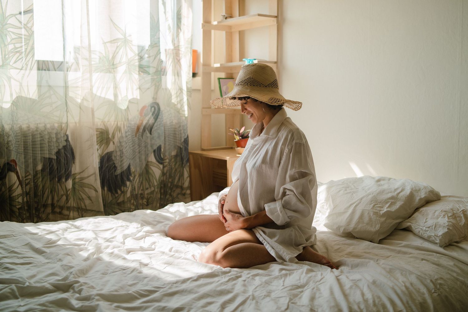 A pregnant woman in a straw hat and linen shirt sits on a white bed, cradling her belly in soft natural light.