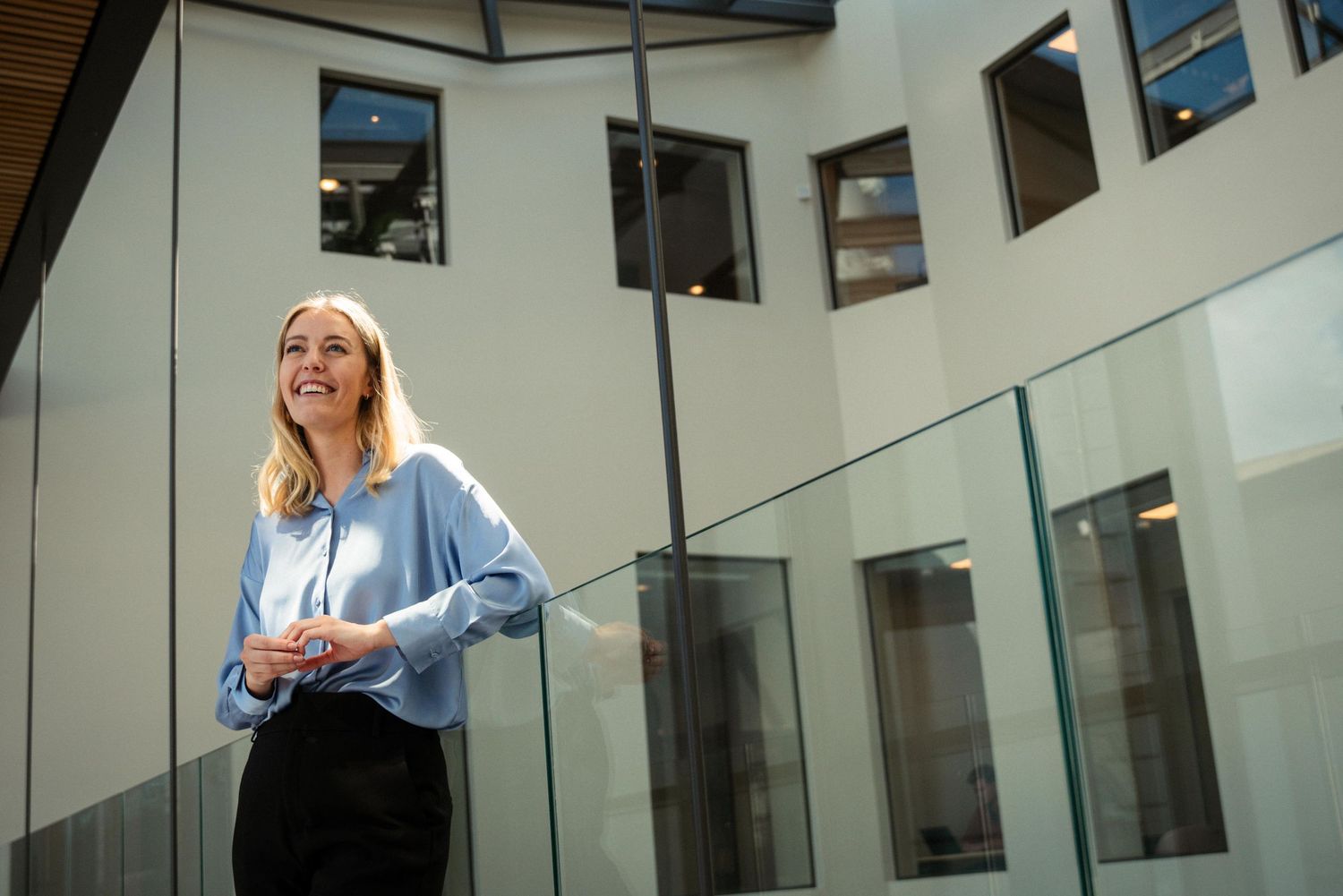 Two women in business attire walk and talk in a modern office hallway with glass walls and natural lighting.