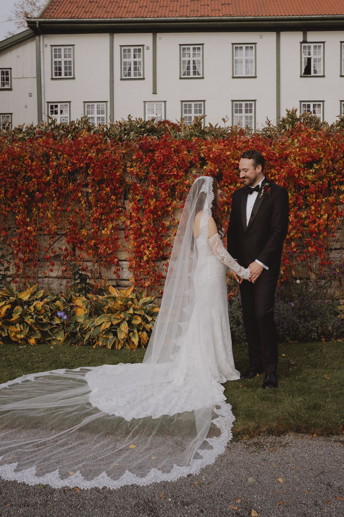 A couple in wedding attire poses against a vibrant red autumn vine-covered wall at a historic building.