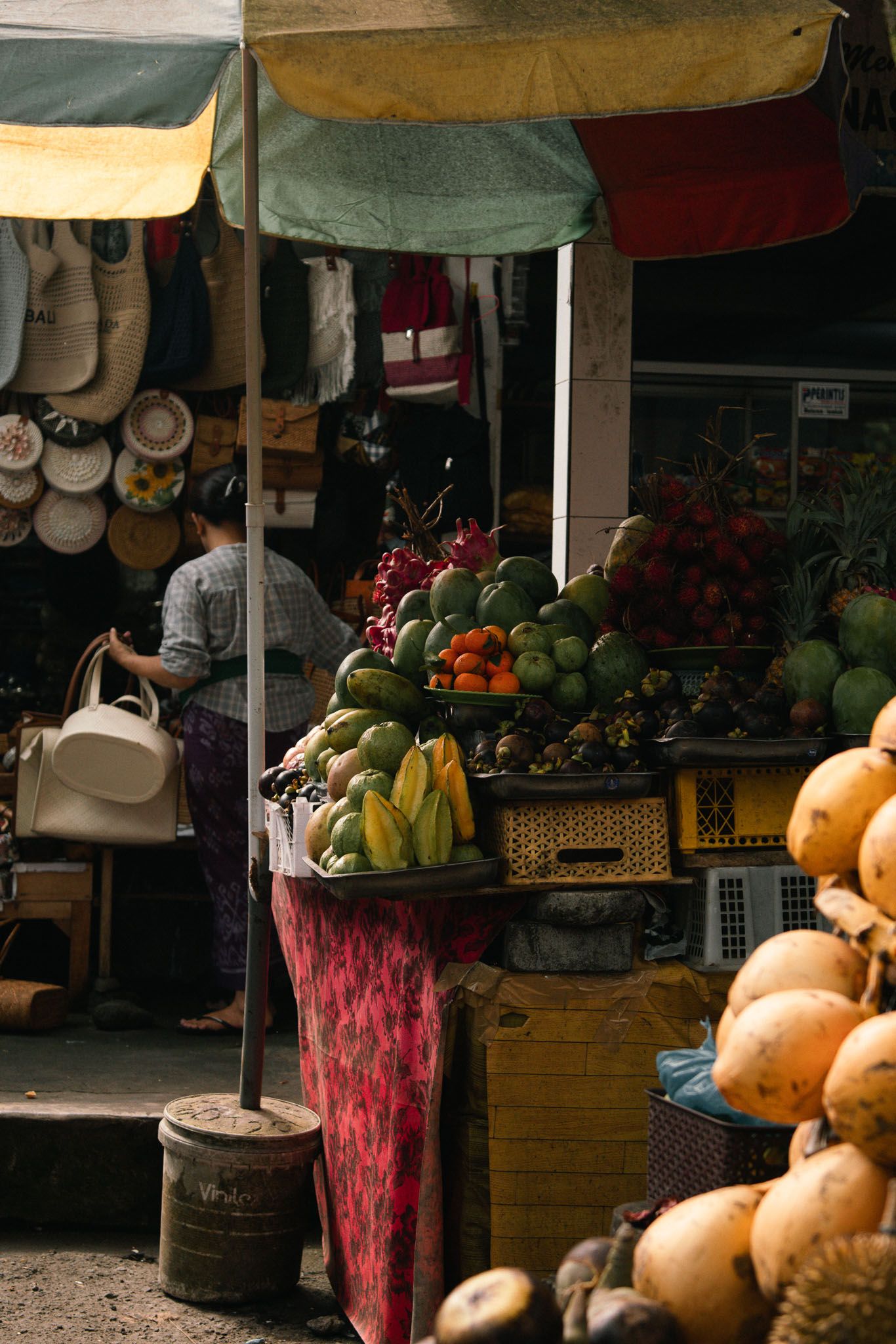 A rustic outdoor market stall displays fresh produce, pumpkins, and flowers with wooden barrels and hanging decor in Bali.