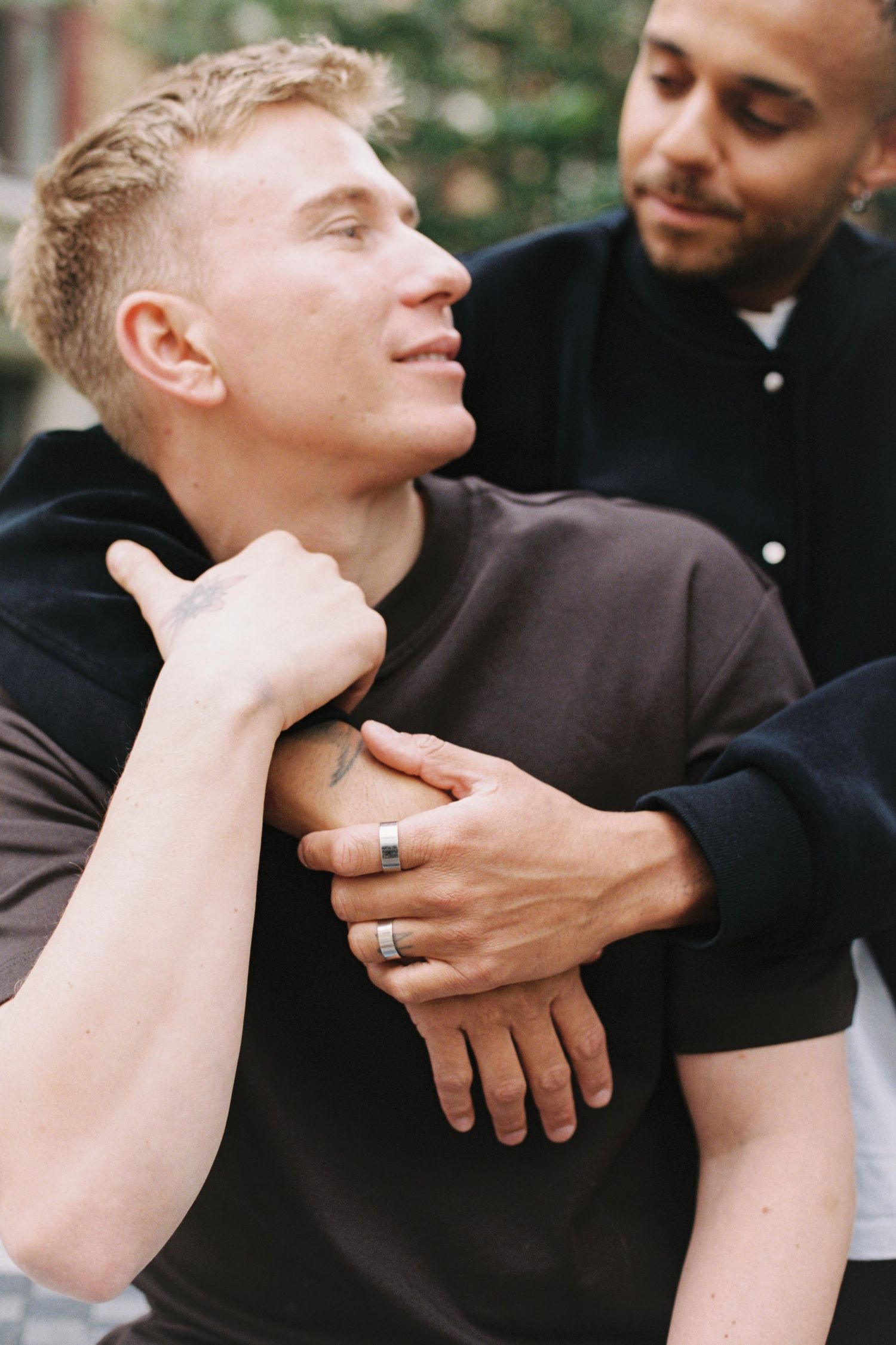Close up portrait of two people embracing outdoors, wearing black shirts and showing their rings.
