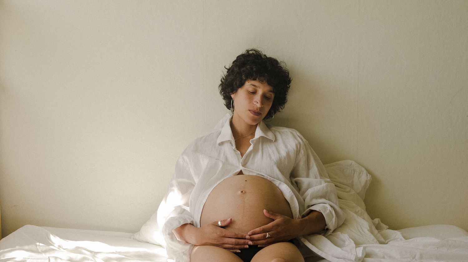 A person wearing a white button-up shirt lies on white bedding while cradling their pregnant belly.