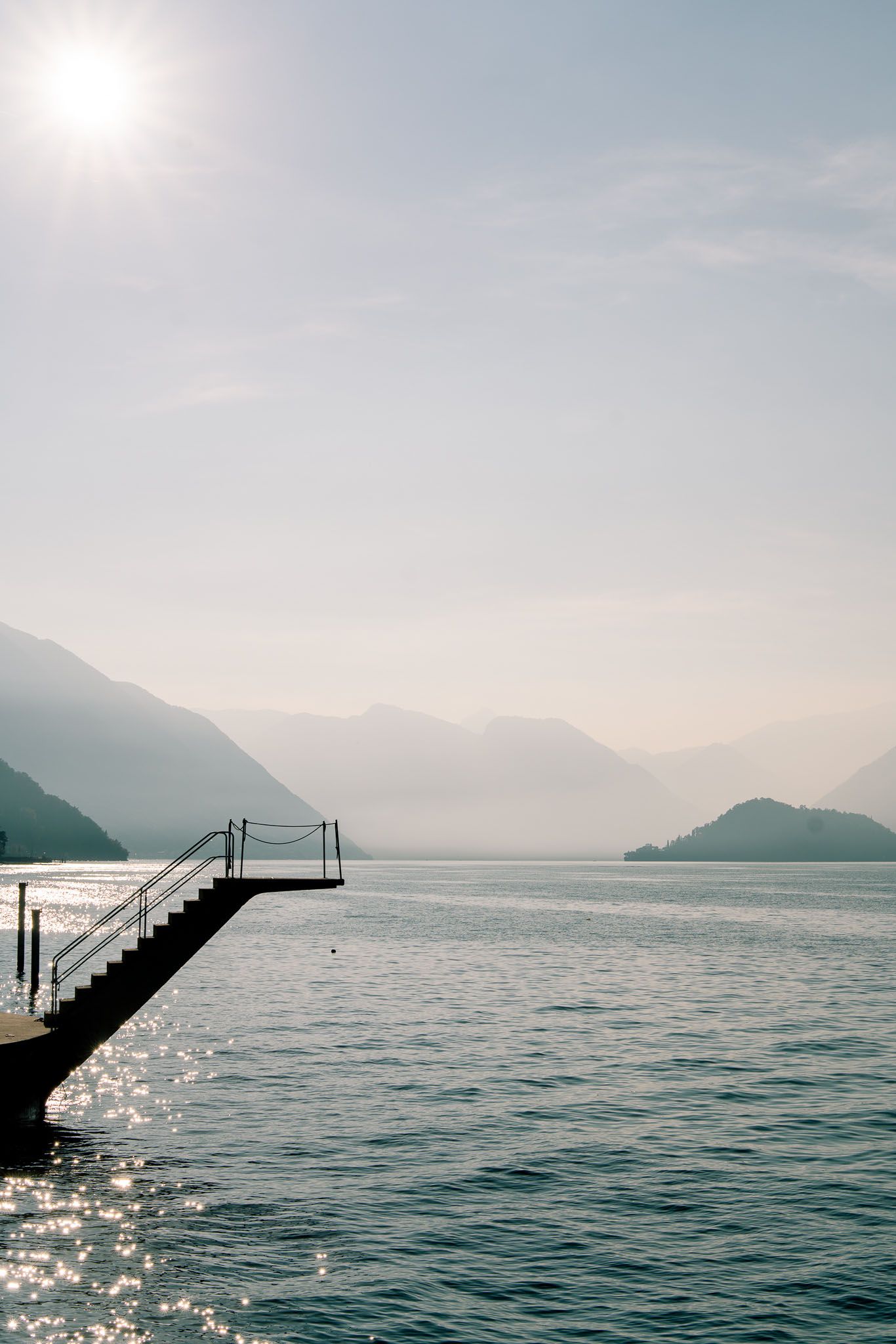 A diving board extends over the sparkling blue waters of Lake Como on a misty summer day.