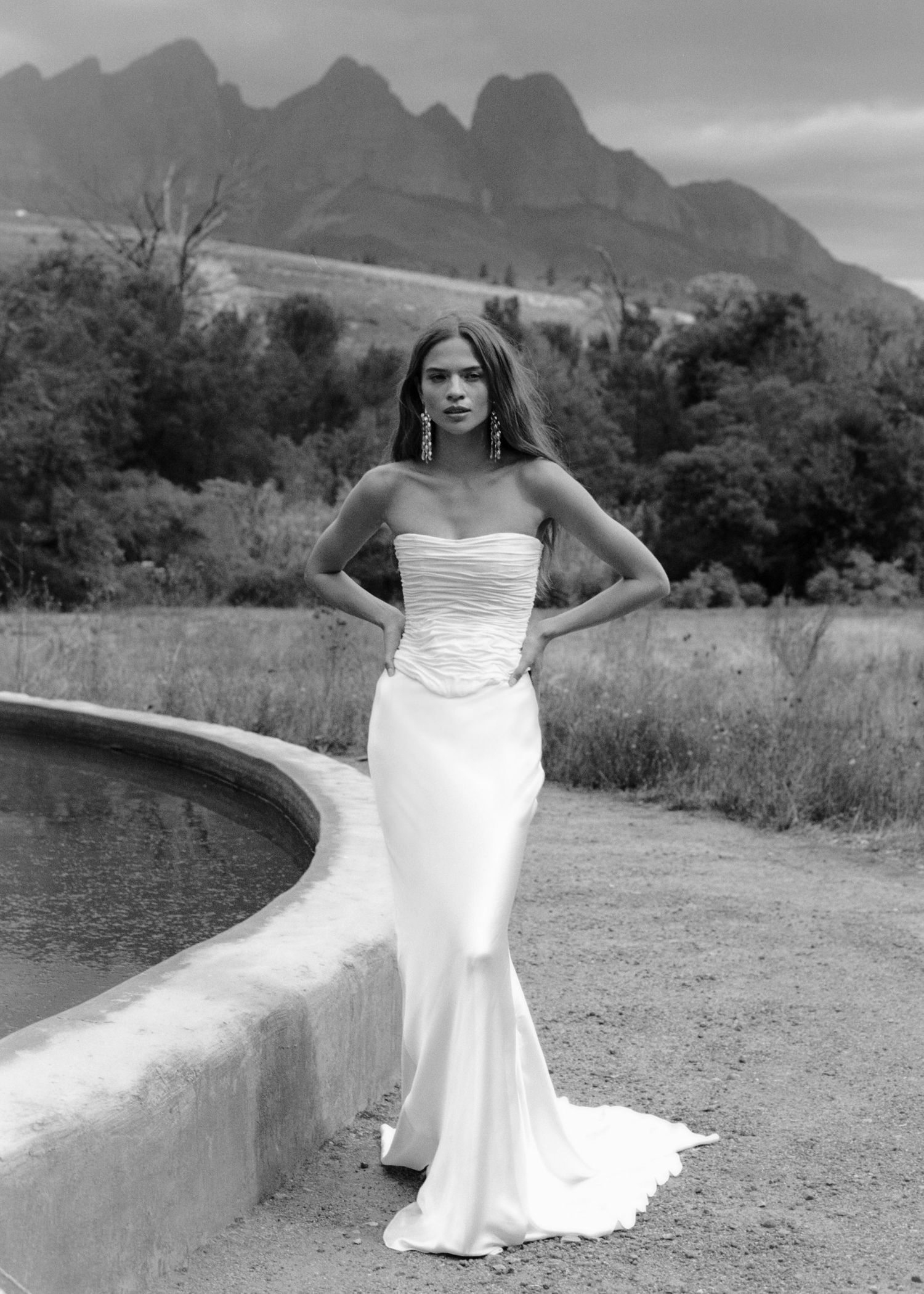 Model in strapless white mermaid wedding gown poses near pool with dramatic mountain backdrop in black and white.
