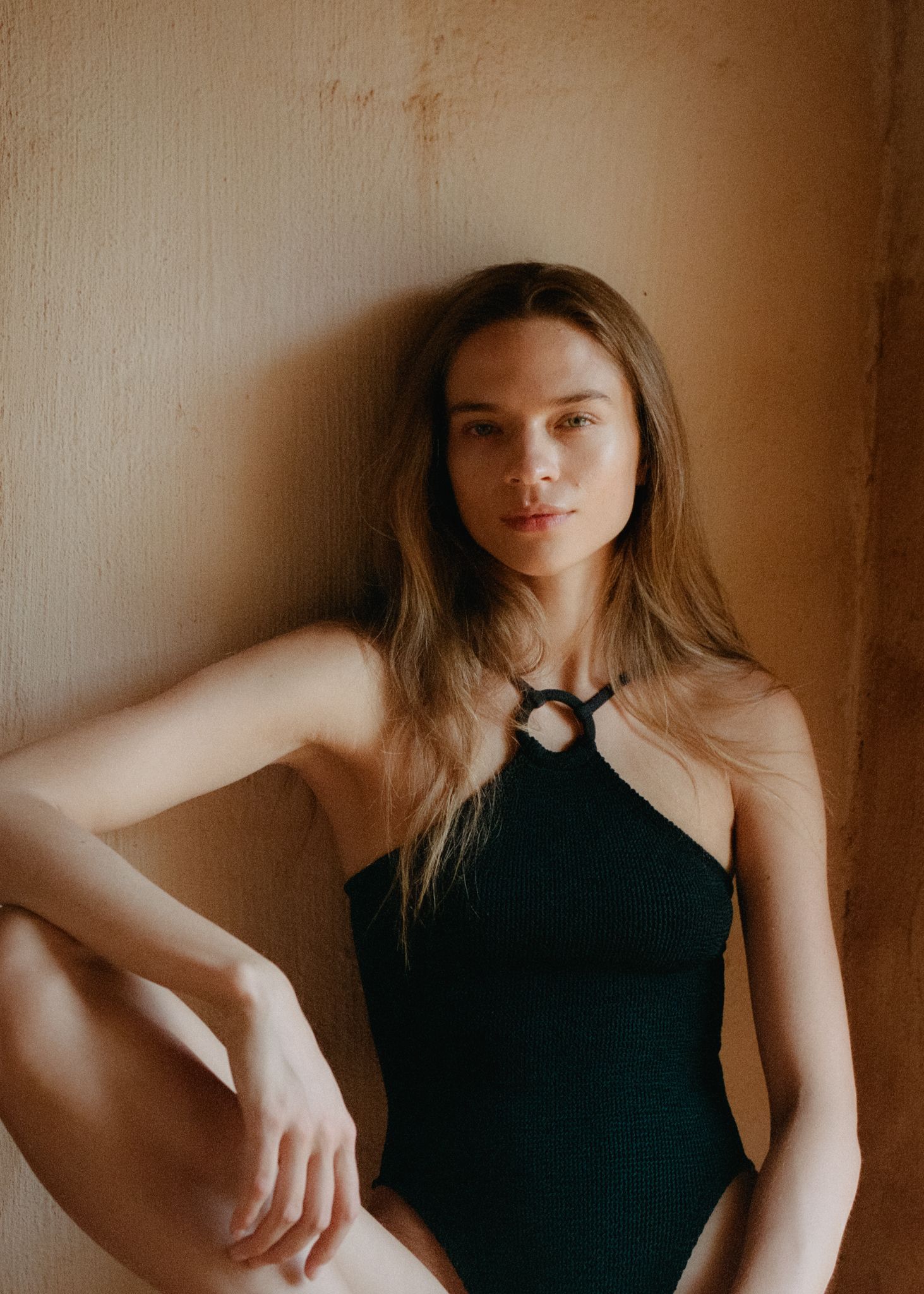 Young woman in black halter swimsuit poses against warm beige wall, looking directly at camera.