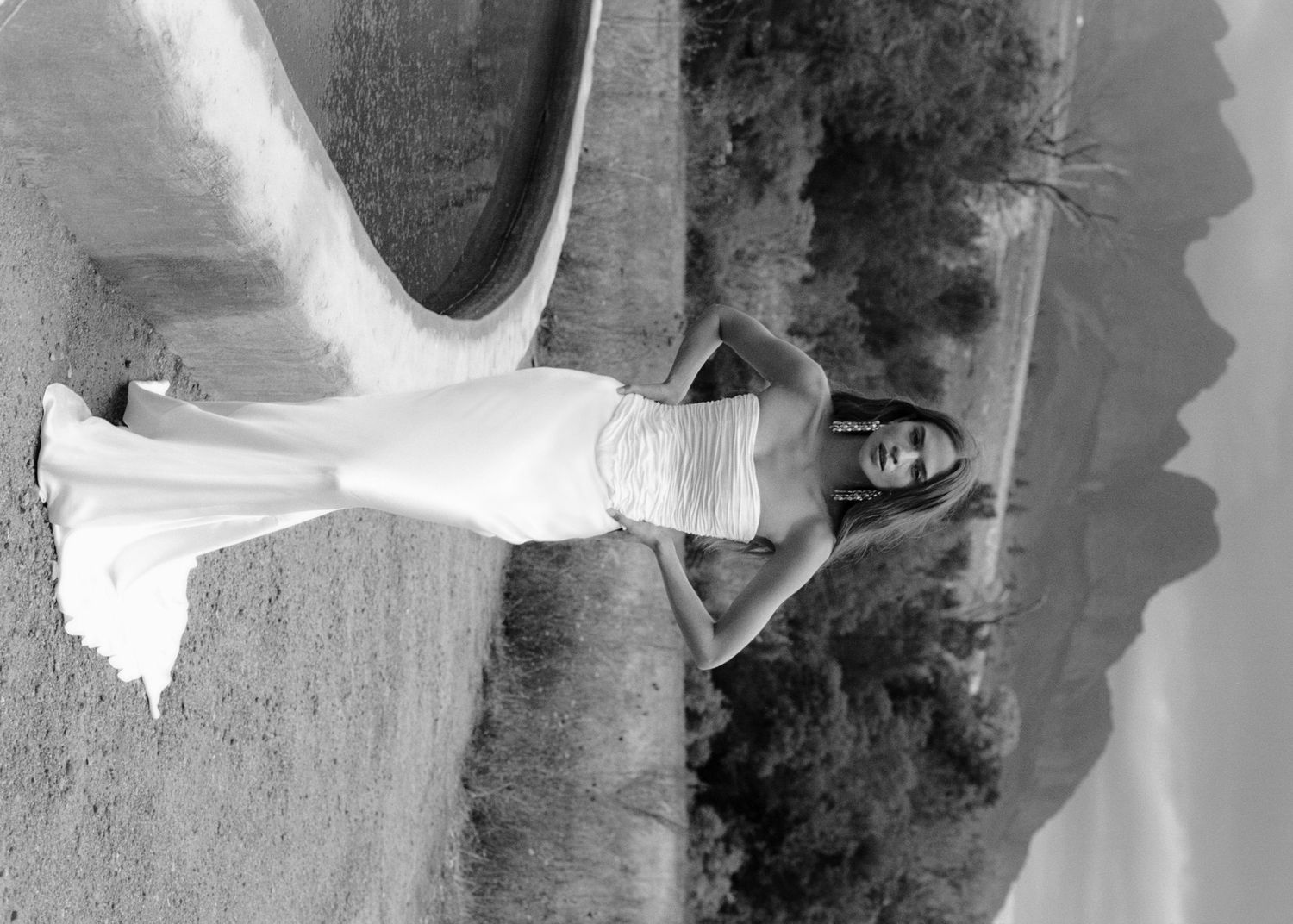 Black and white photo of a woman in a white wedding gown posing outdoors near a curved wall with hills in the background.