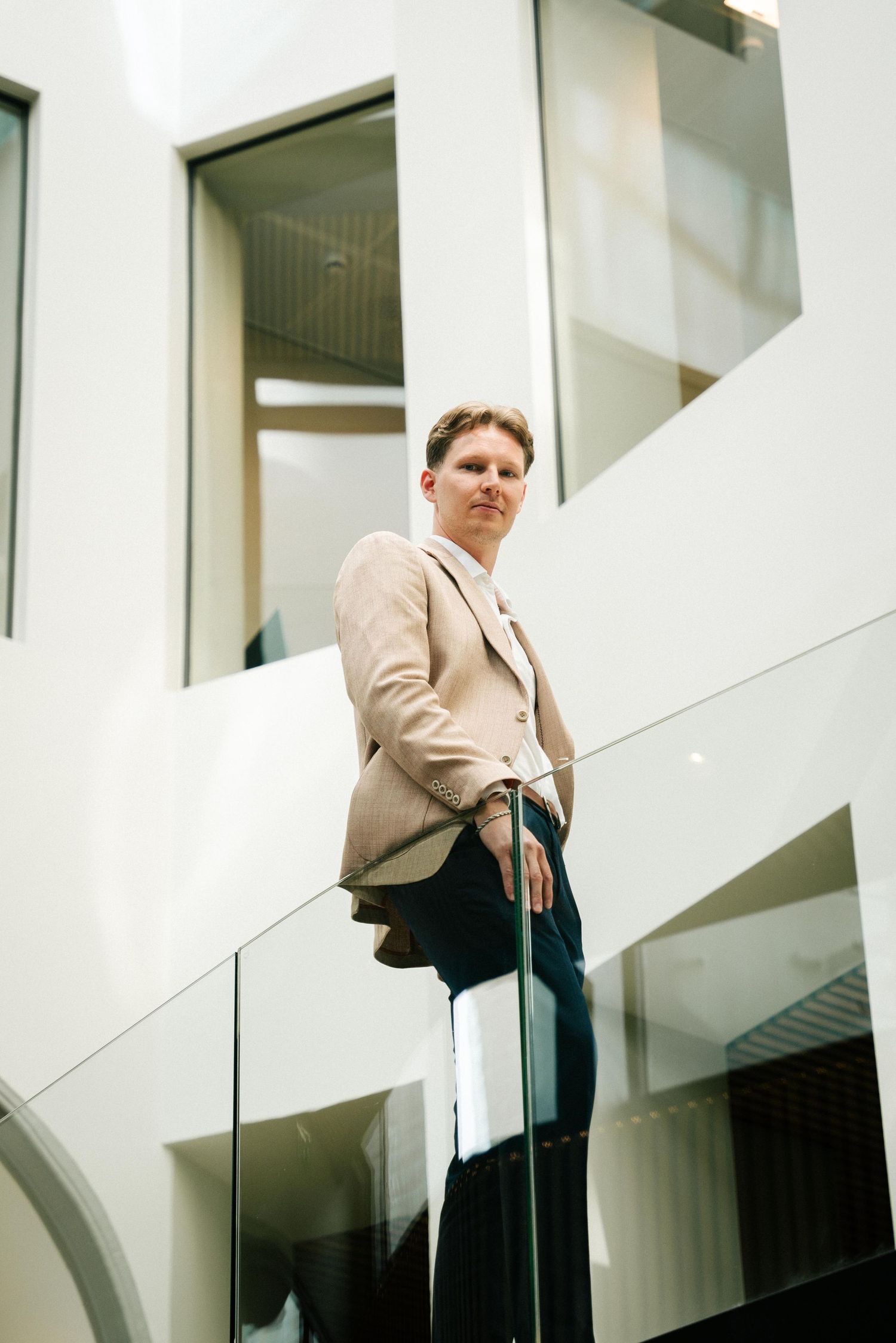 Person in beige blazer standing against modern white interior architecture with glass railings and curved archway.
