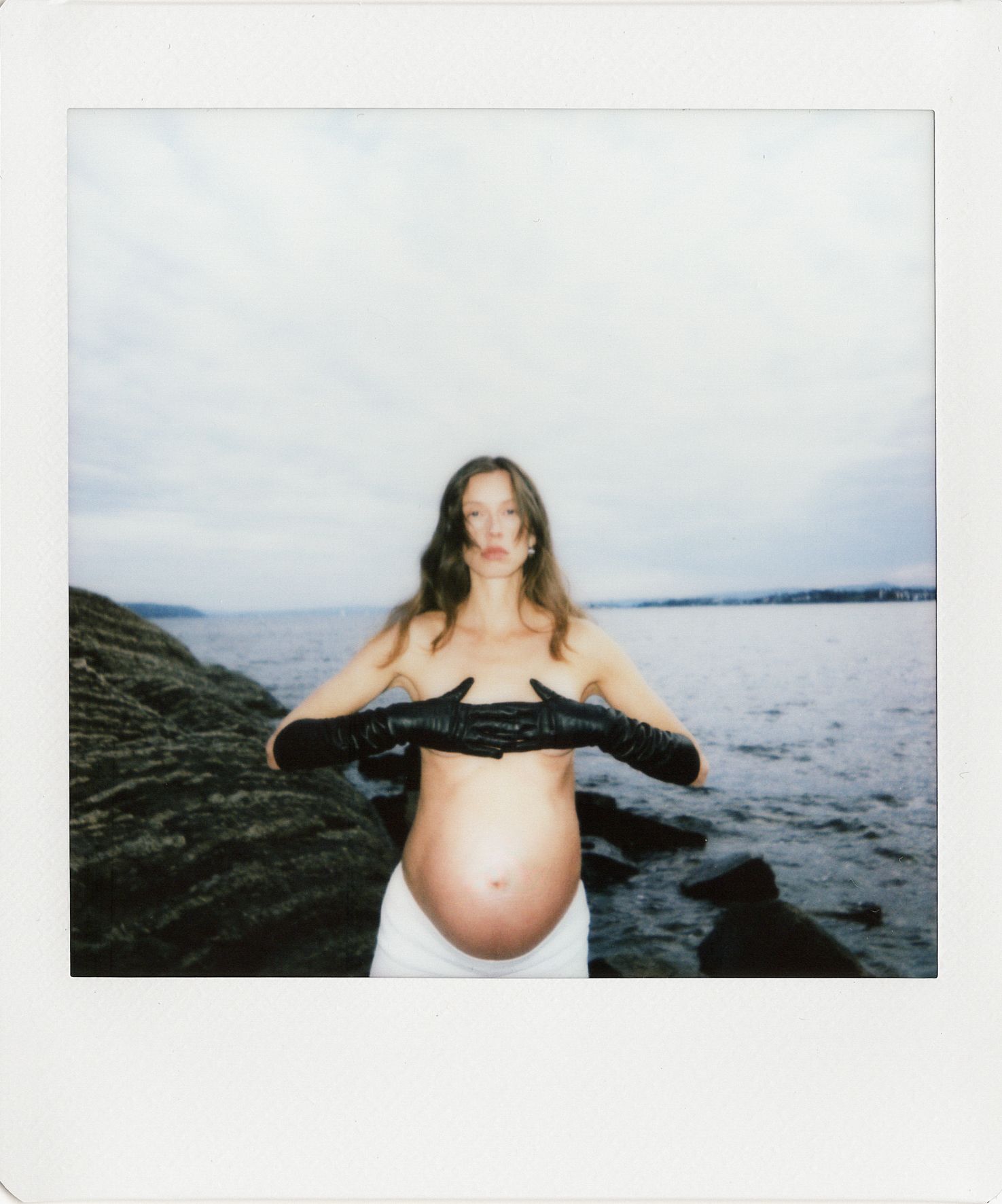 Pregnant woman in black leather gloves posing topless by rocky seaside in a Polaroid-style photo.