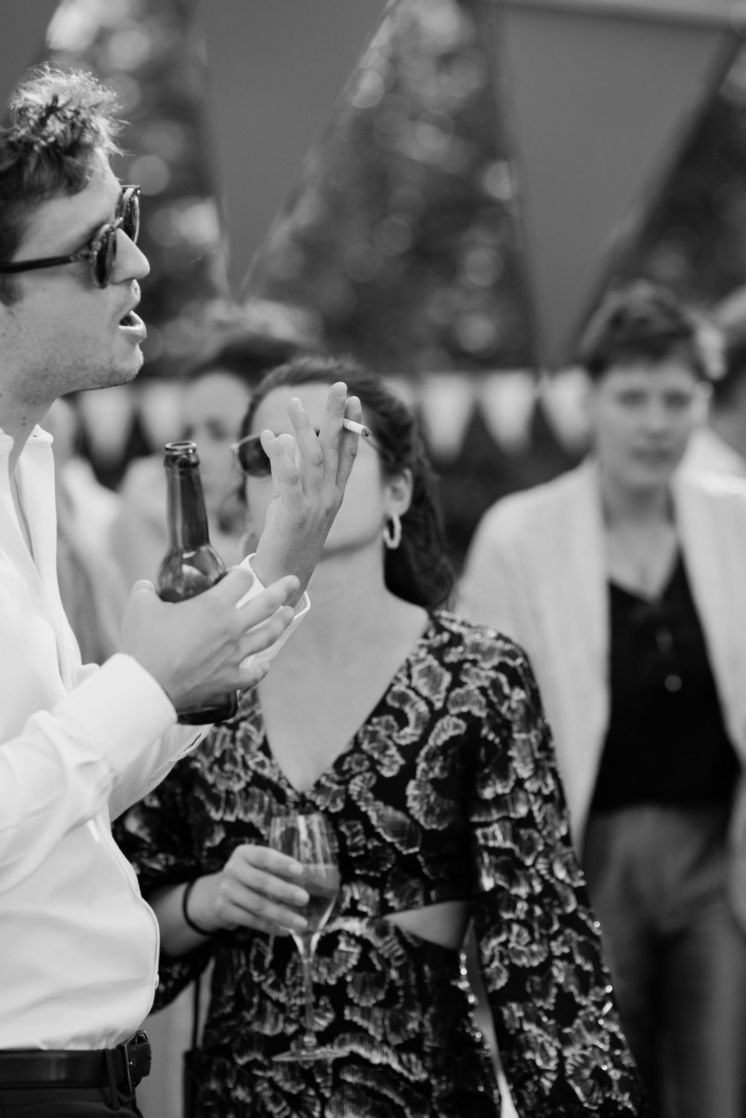 Black and white photo of man with sunglasses holding beer bottle at outdoor party with guests in background.