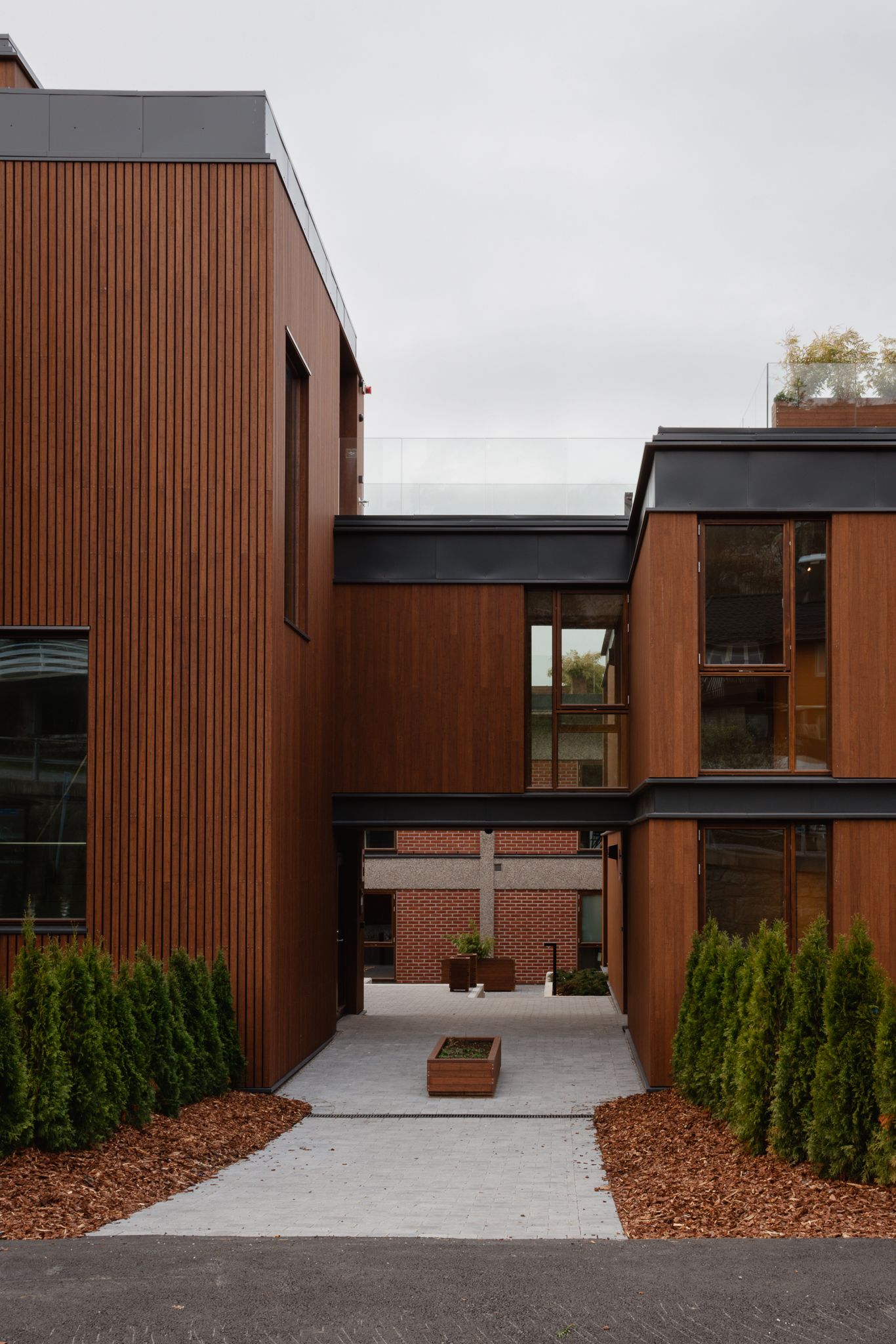 Modern building exterior with rust-colored metal panels and concrete walkway featuring green shrubs and mulch landscaping.