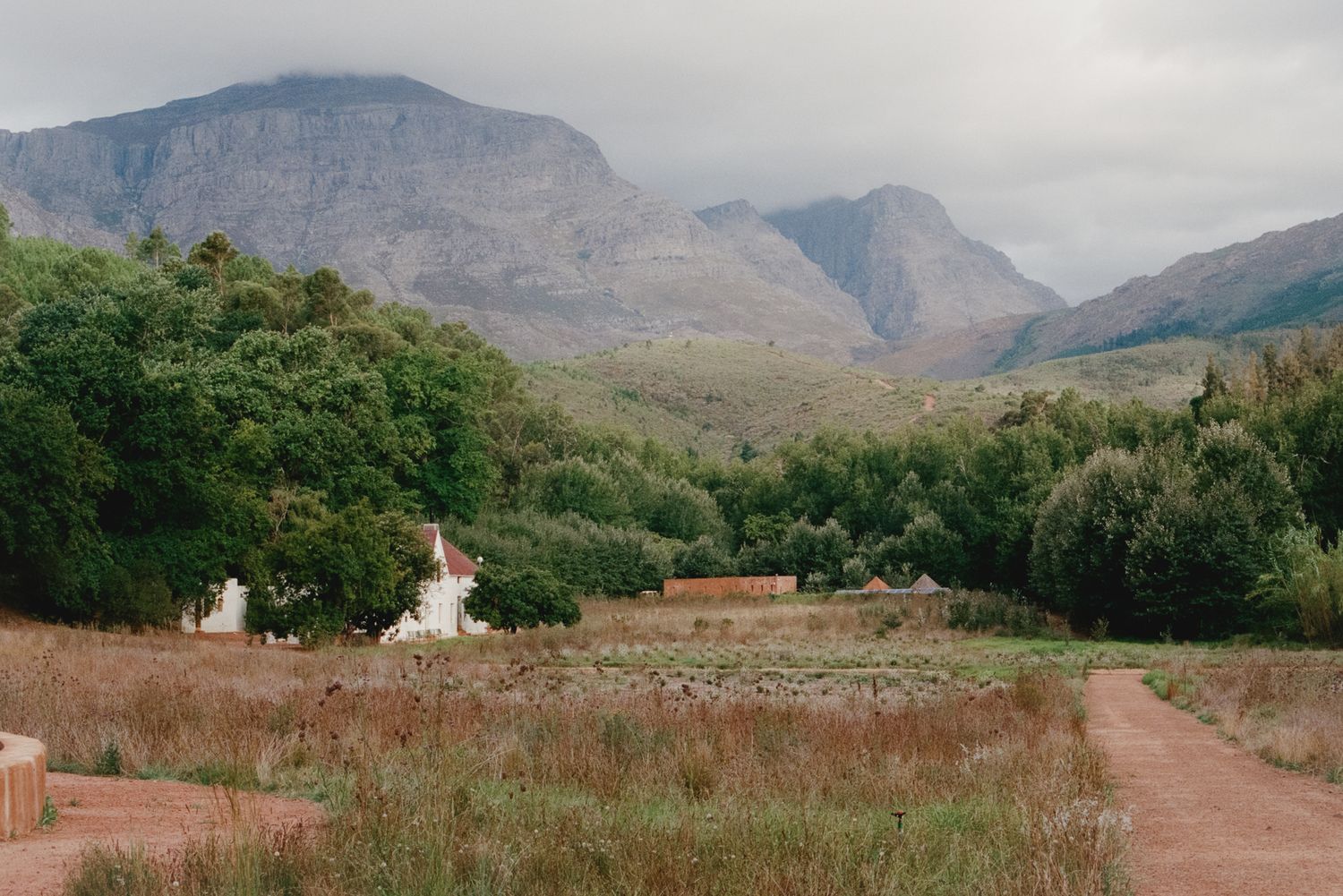 Rural landscape with white farmhouse nestled among green trees, dry grasslands, and dramatic misty mountains in background.